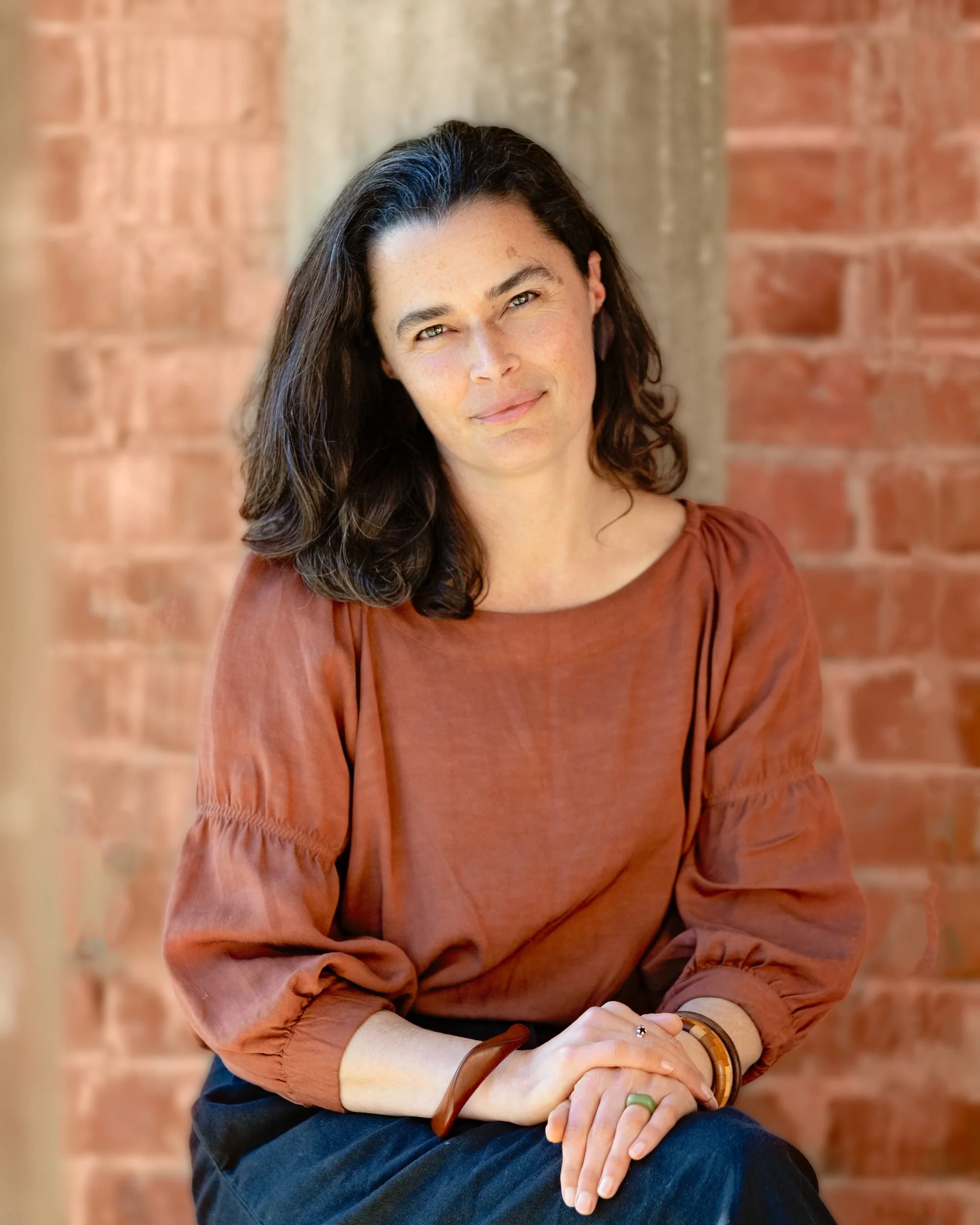 A woman with dark, wavy hair and light skin sits against a brick wall, wearing a rust-colored top and accessorizing with bracelets and a ring.