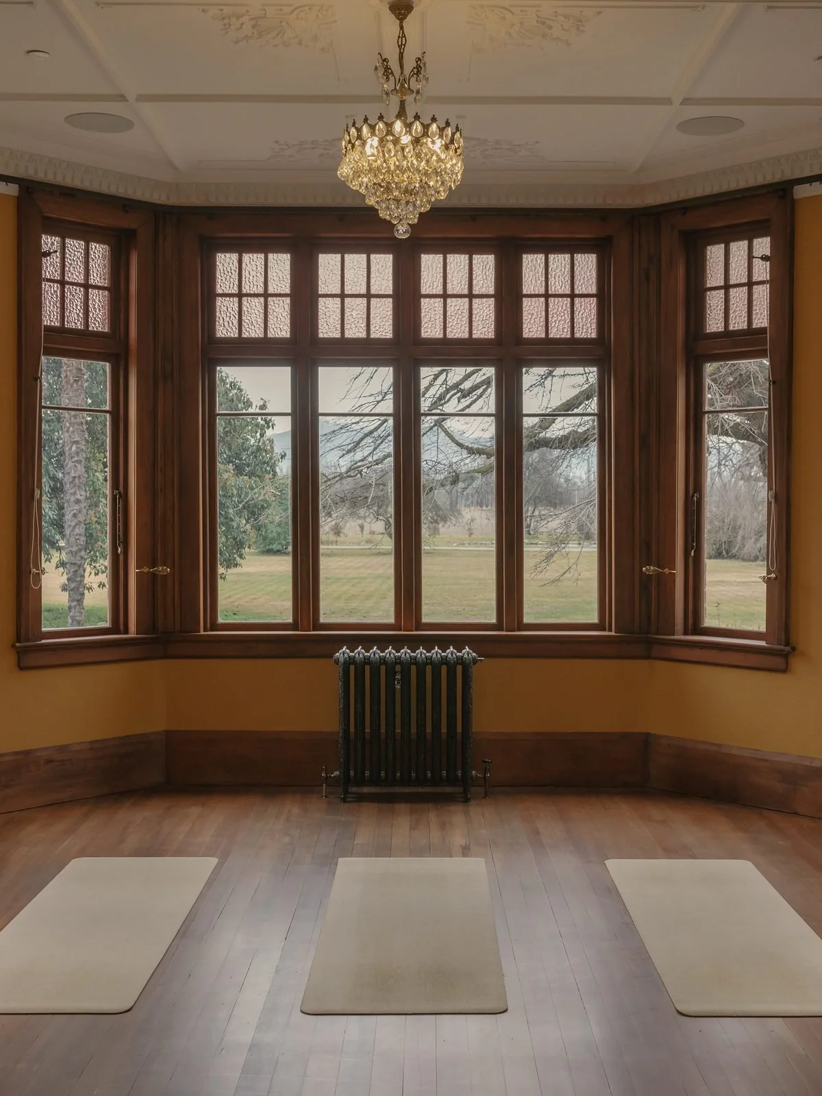 A spacious room with a wooden floor, three meditation mats, large windows, and a chandelier hanging from a decorated ceiling.