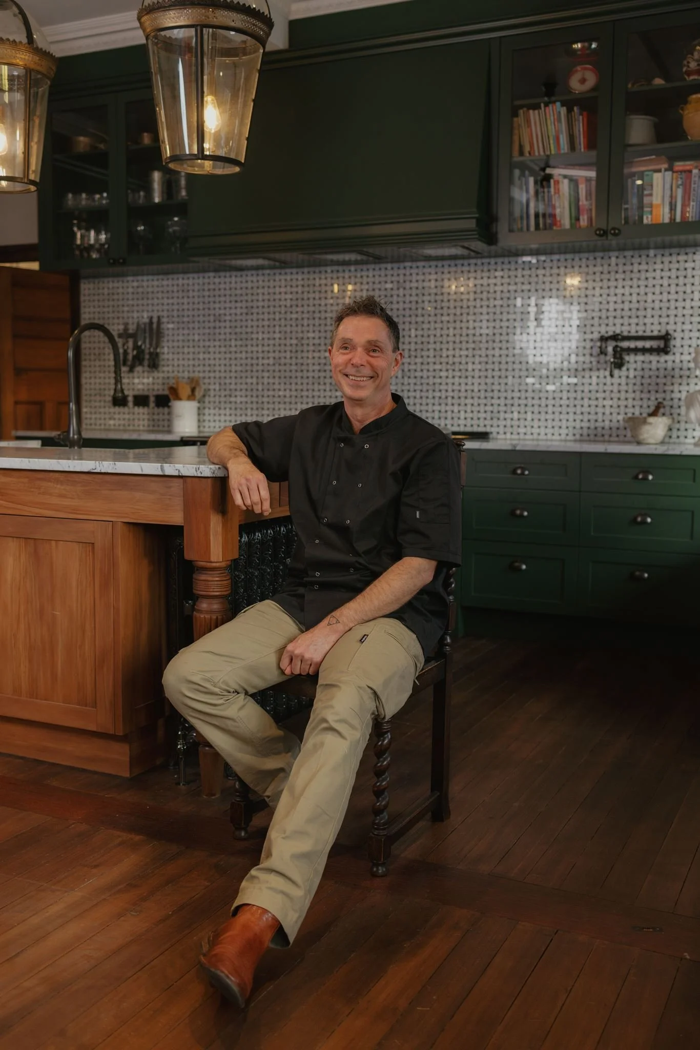 A man in a black chef jacket and light-colored pants sitting on a wooden chair at a kitchen island, smiling. The kitchen has dark green cabinets, a tiled backsplash, and a bookshelf with cookbooks and decorative items. There are two large hanging lights above the man.