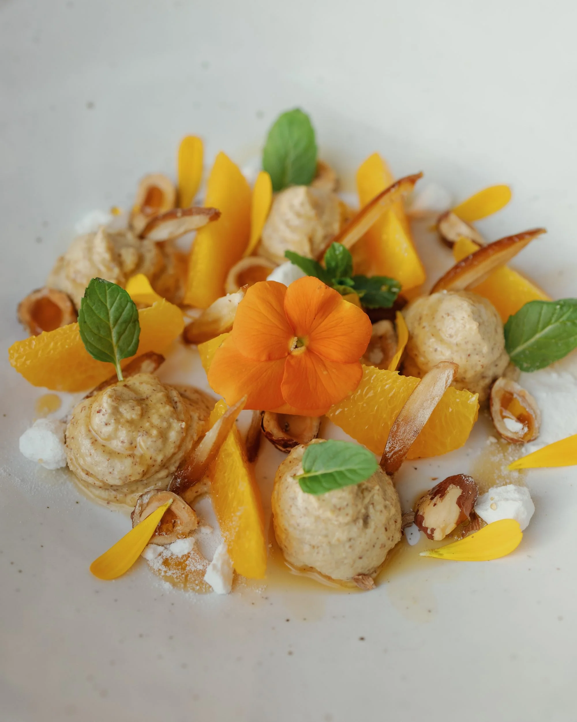Dessert featuring orange segments, edible flowers, green mint leaves, toasted coconut flakes, and dollops of cream or mousse on a white plate.