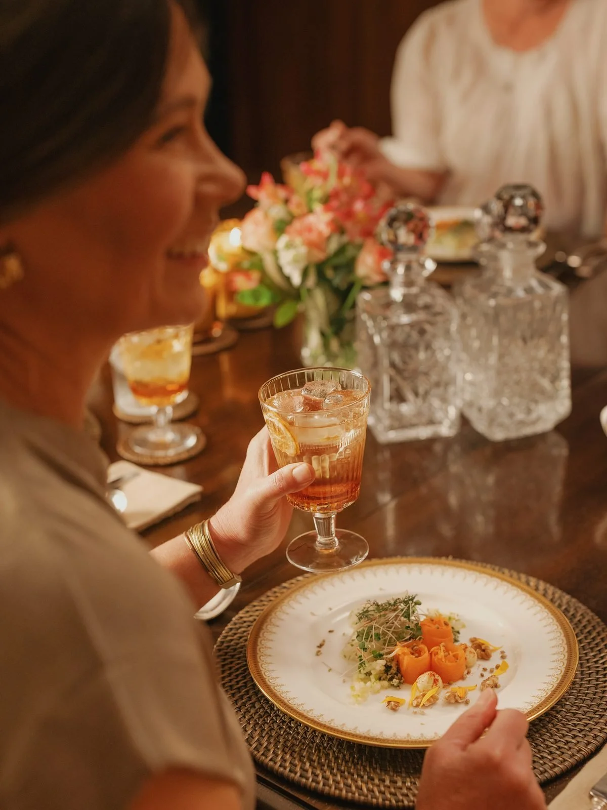 A woman holding a glass of iced tea or cocktail at a formal dinner table, with a plated gourmet meal, floral centerpiece, and decorative glass bottles in the background.