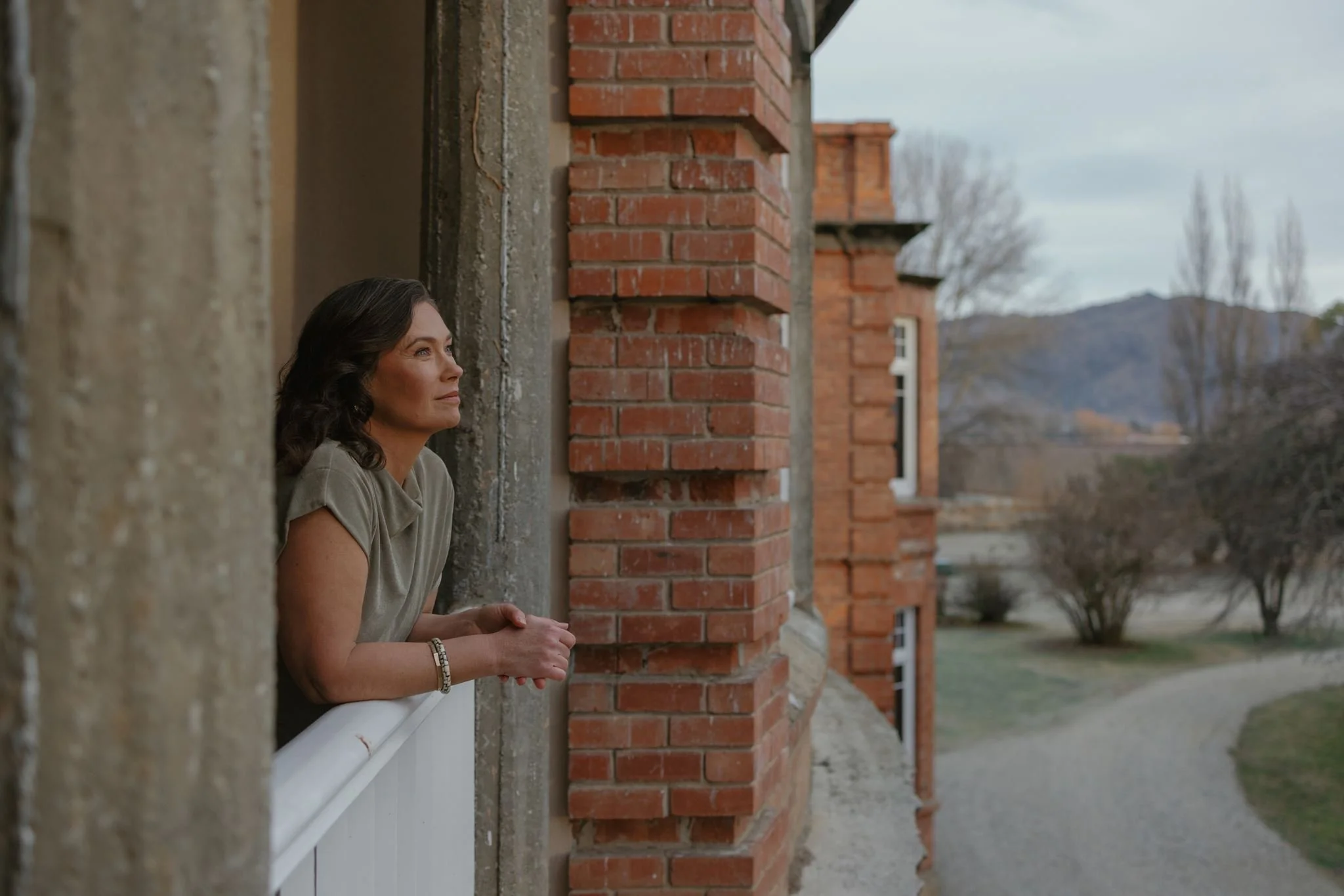 A woman with dark hair and wearing a gray shirt sits on a porch, leaning against a brick pillar, looking into the distance with a thoughtful expression.