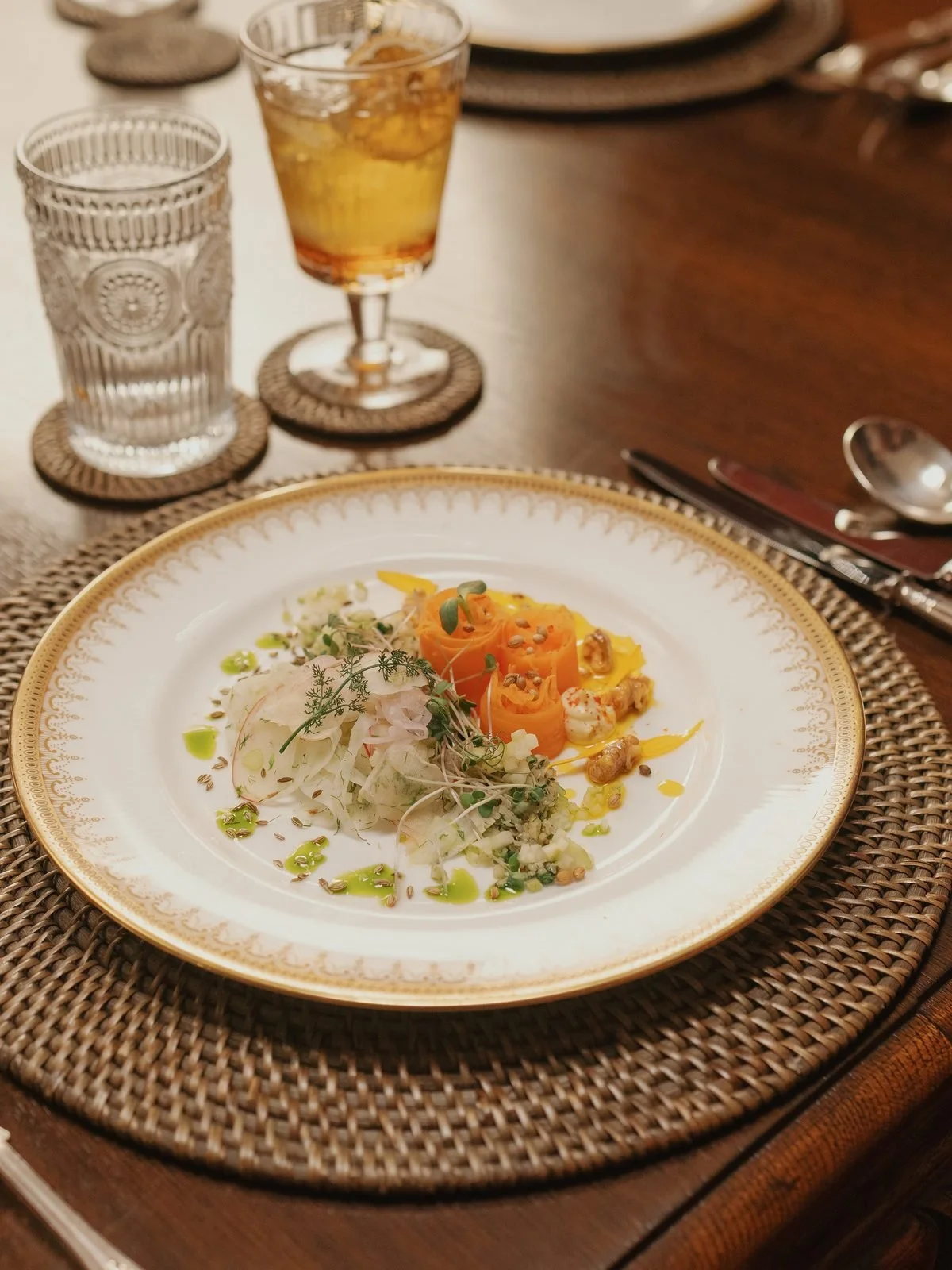 Elegant dinner table with a white and gold-rimmed plate of gourmet food, including shredded vegetables, seeds, and a rolled vegetable garnish, garnished with herbs and oil. Two glasses, one with water and the other with a cocktail, sit behind the plate. Silver utensils rest on a napkin beside the plate, and the table has woven placemats.