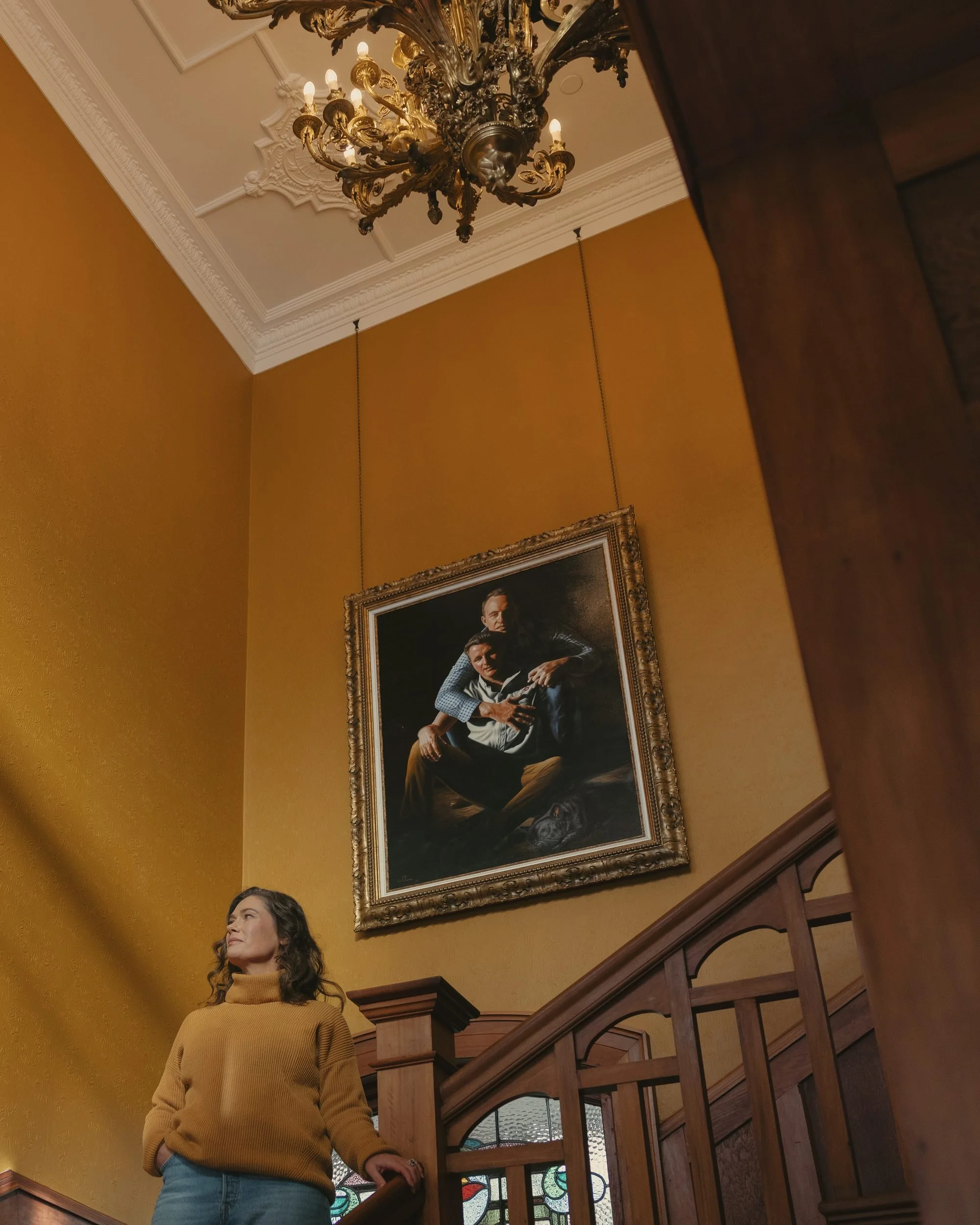 A woman in a yellow turtleneck sweater standing on a staircase in an elegant room with a gold framed portrait painting on a yellow wall and an ornate chandelier hanging from the ceiling.