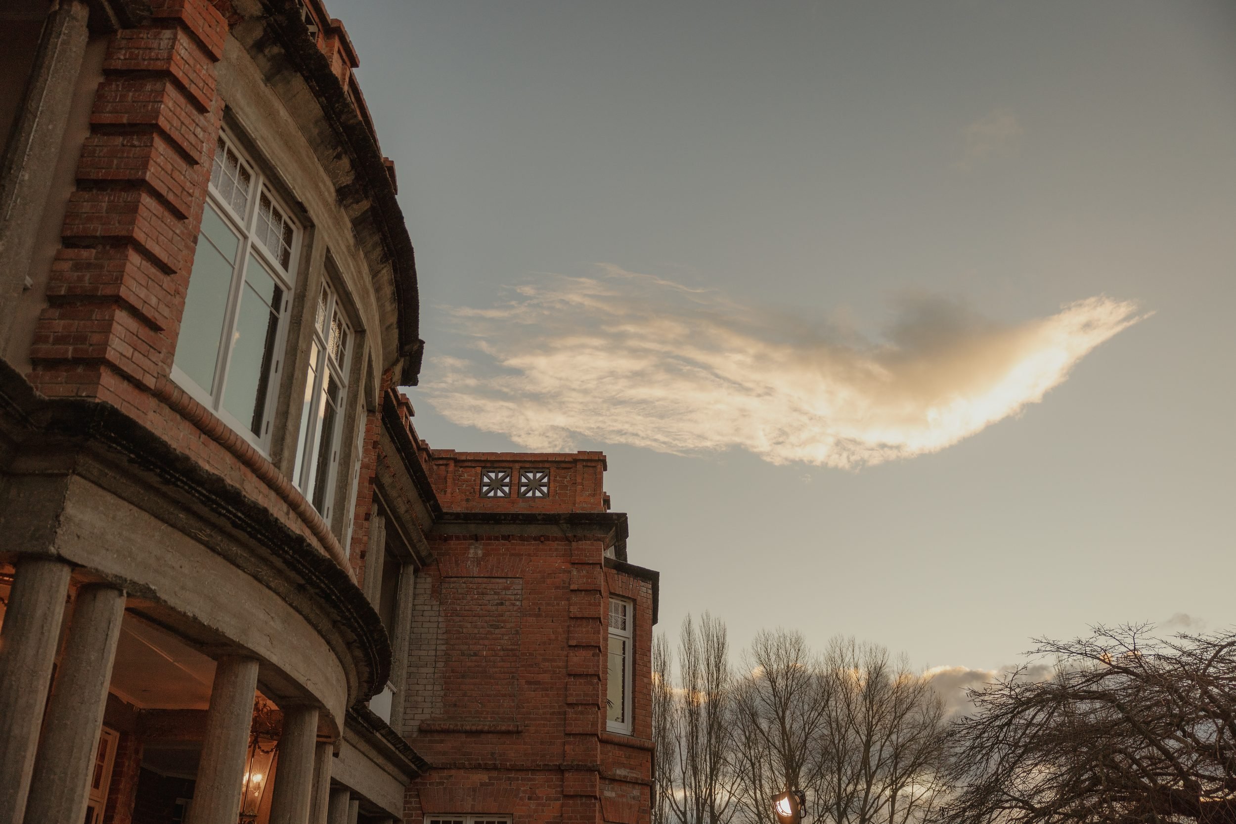 The photo shows the side of a brick house with white-framed windows and a rounded architectural feature supported by columns. The sky has a mix of clouds and a setting or rising sun, creating a warm glow. In the background, there are leafless trees.