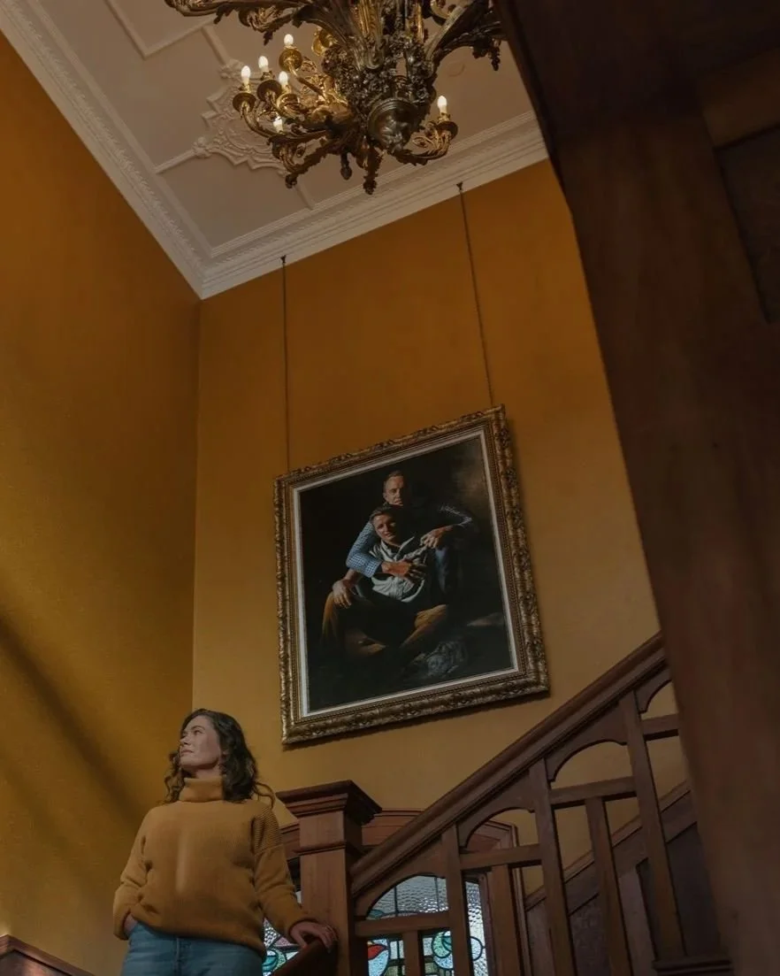 A woman in a tan sweater and jeans standing on a staircase, looking away, inside a room with yellow walls. Above her, a large framed painting of two people sitting close together hangs on the wall, and a chandelier is on the ceiling.