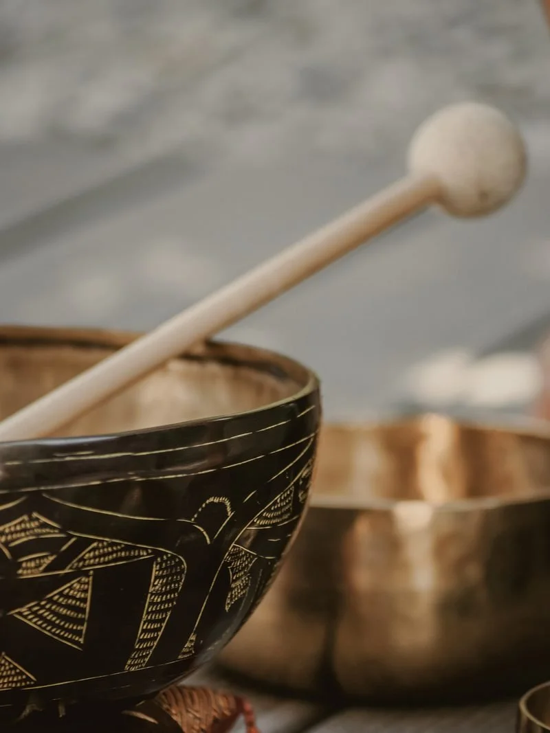 Close-up of a black and gold musical singing bowl with a wooden mallet resting inside, and other metallic bowls visible in the background.