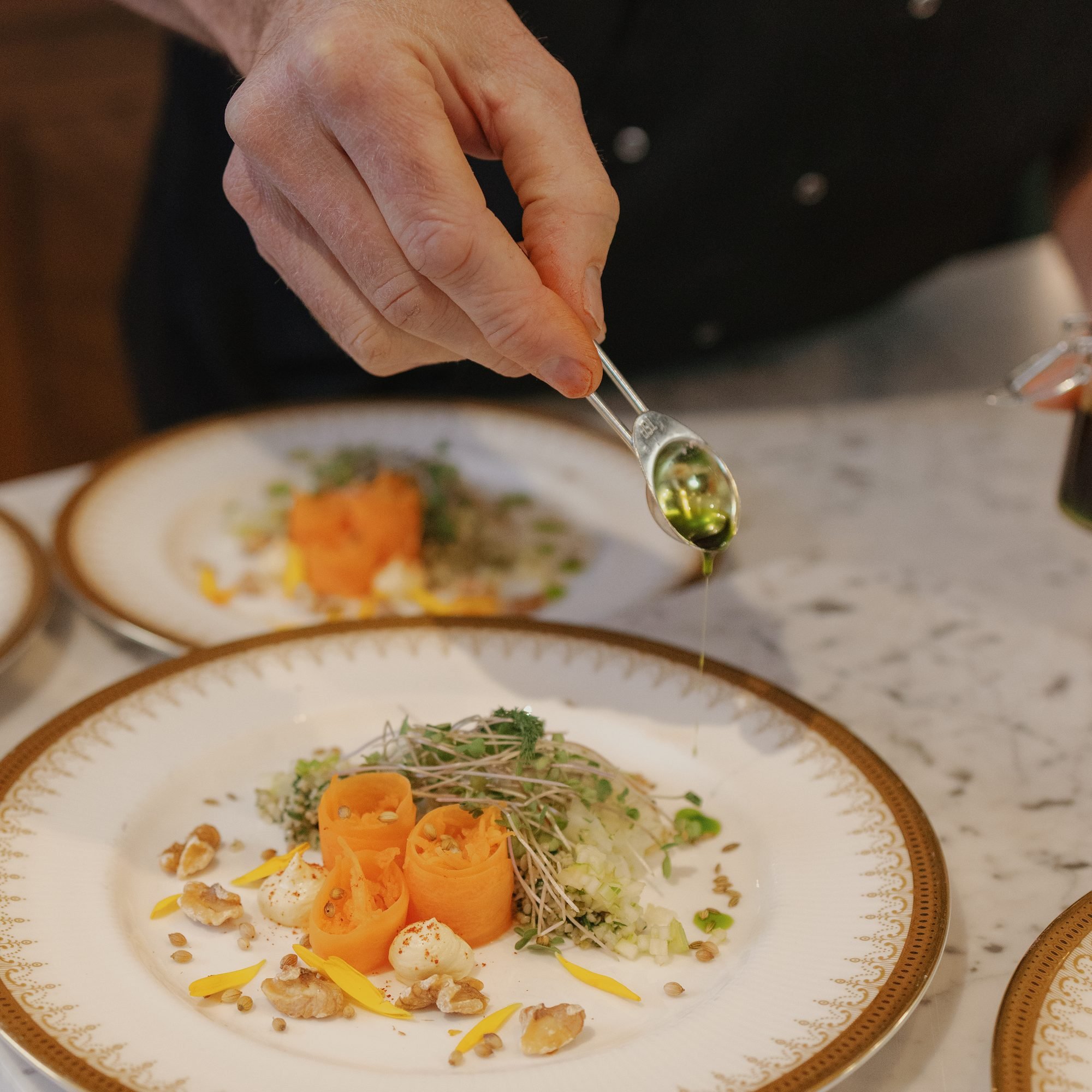A hand drizzling green sauce over a gourmet salad with vegetables, microgreens, and cheese on a decorative white and gold plate.