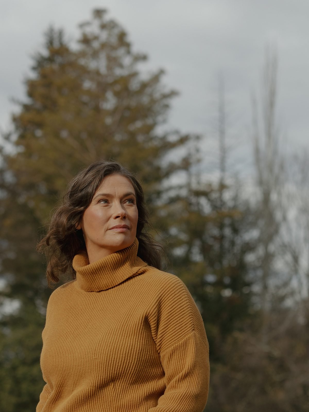 A woman with wavy brown hair wearing a mustard-colored turtleneck sweater stands outdoors with trees in the background on a cloudy day.