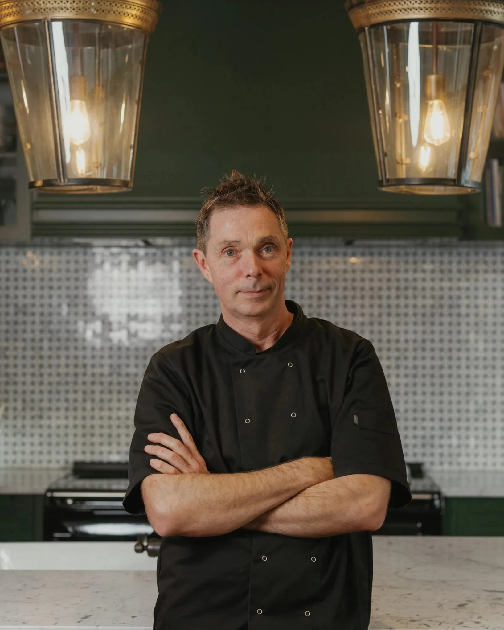 A male chef standing with his arms crossed in a kitchen, with two large pendant lights hanging overhead and a stove in the background.