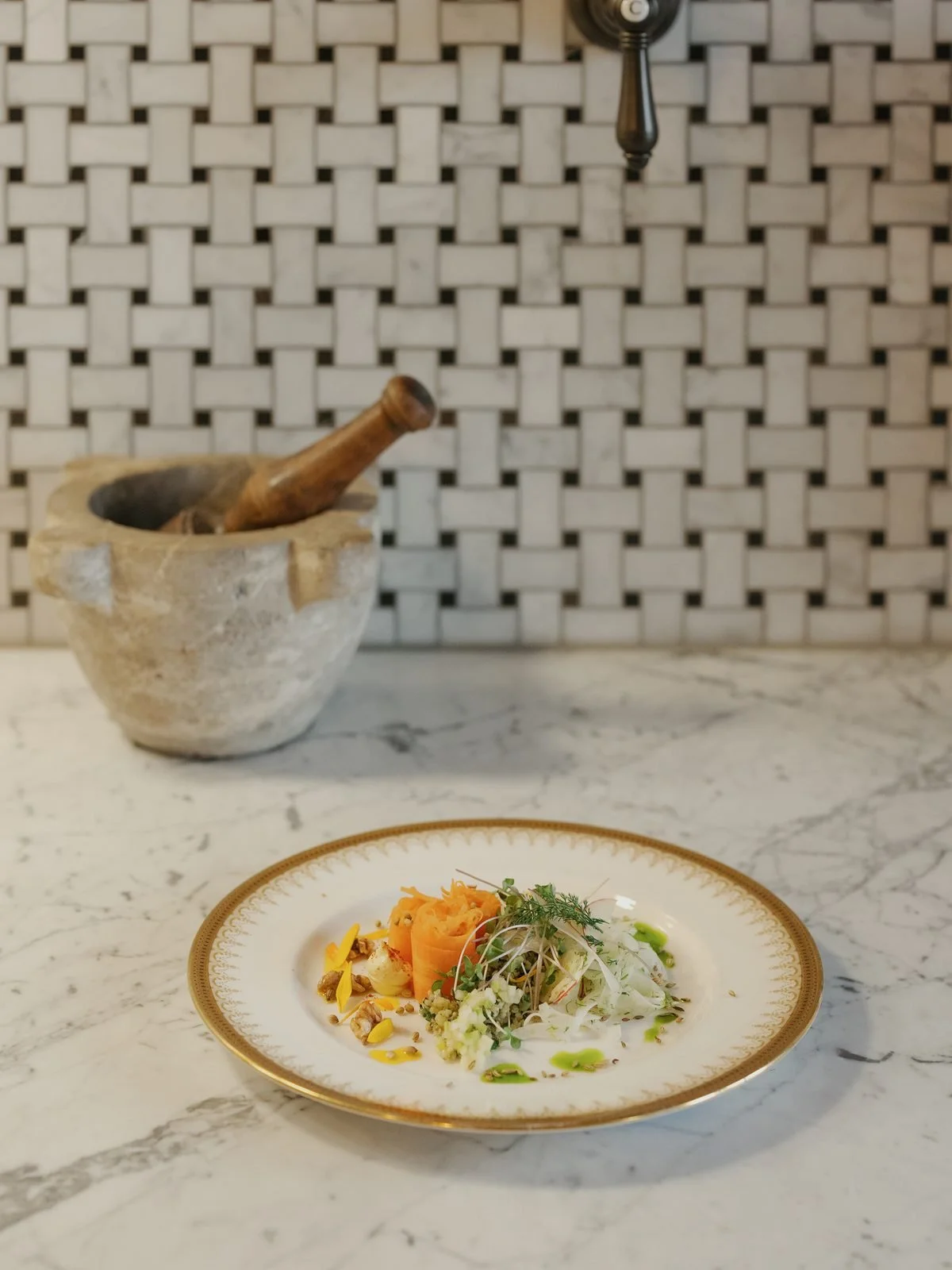 A marble countertop with a decorative plate of salad, including sliced radish, carrot, and greens, in the foreground. In the background, there is a stone mortar with a wooden pestle inside, and a woven backsplash. A hand-crank coffee or spice grinder is mounted on the wall above.