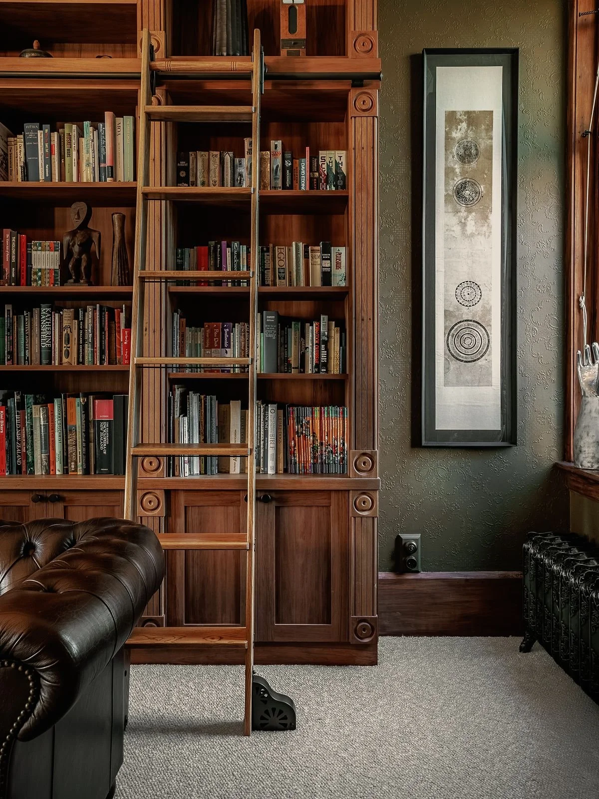 A wooden bookshelf with a built-in ladder against a textured green wall, decorated with books and sculptures. Part of a leather armchair and a decorative vase on a side table are visible.