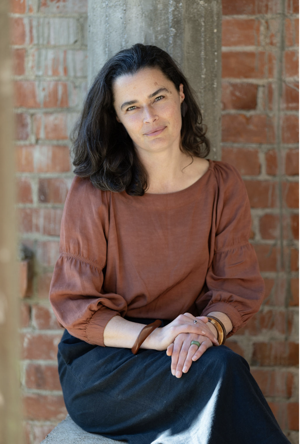 A woman with dark, wavy hair, wearing a rust-colored blouse with puffed sleeves, sitting against a brick wall, looking at the camera with a slight smile.