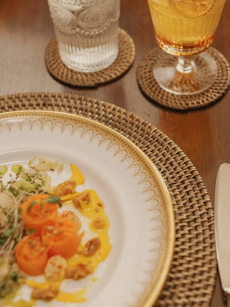 Part of a fancy dinner table setting with a decorative white and gold rimmed plate holding food, a glass of whiskey or dark liquor, and a glass of water, all on woven coasters and a wooden table.