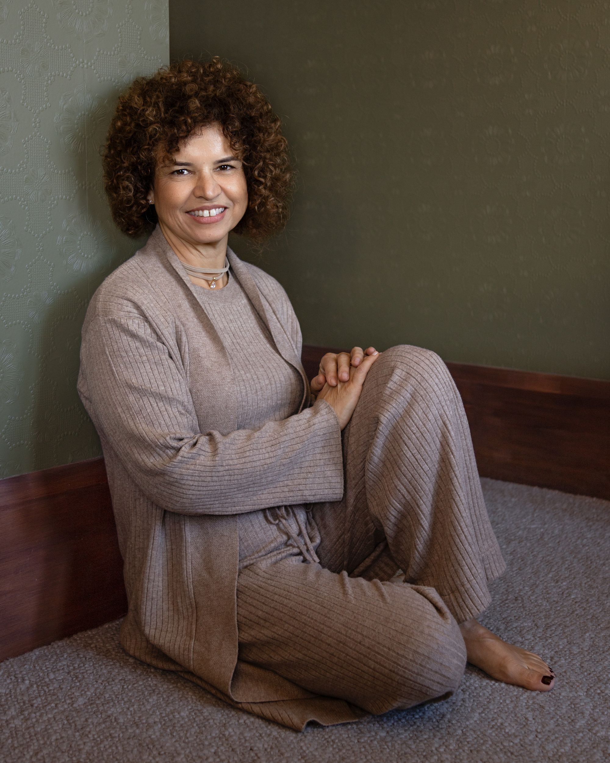 A woman with curly brown hair wearing beige clothing sitting on a carpeted floor with her hands clasped and smiling at the camera.