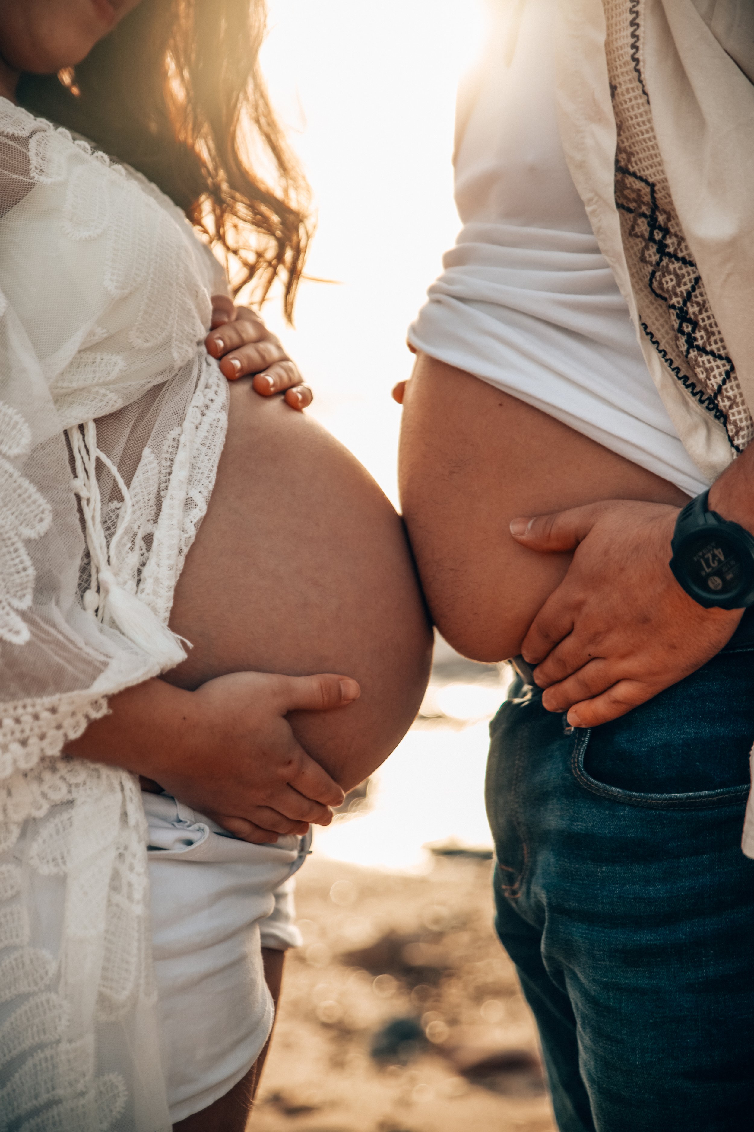 Close-up of a pregnant woman and a man touching her belly, with the woman wearing a lace top and the man wearing a beige shirt with a pattern, both standing outdoors at sunset.