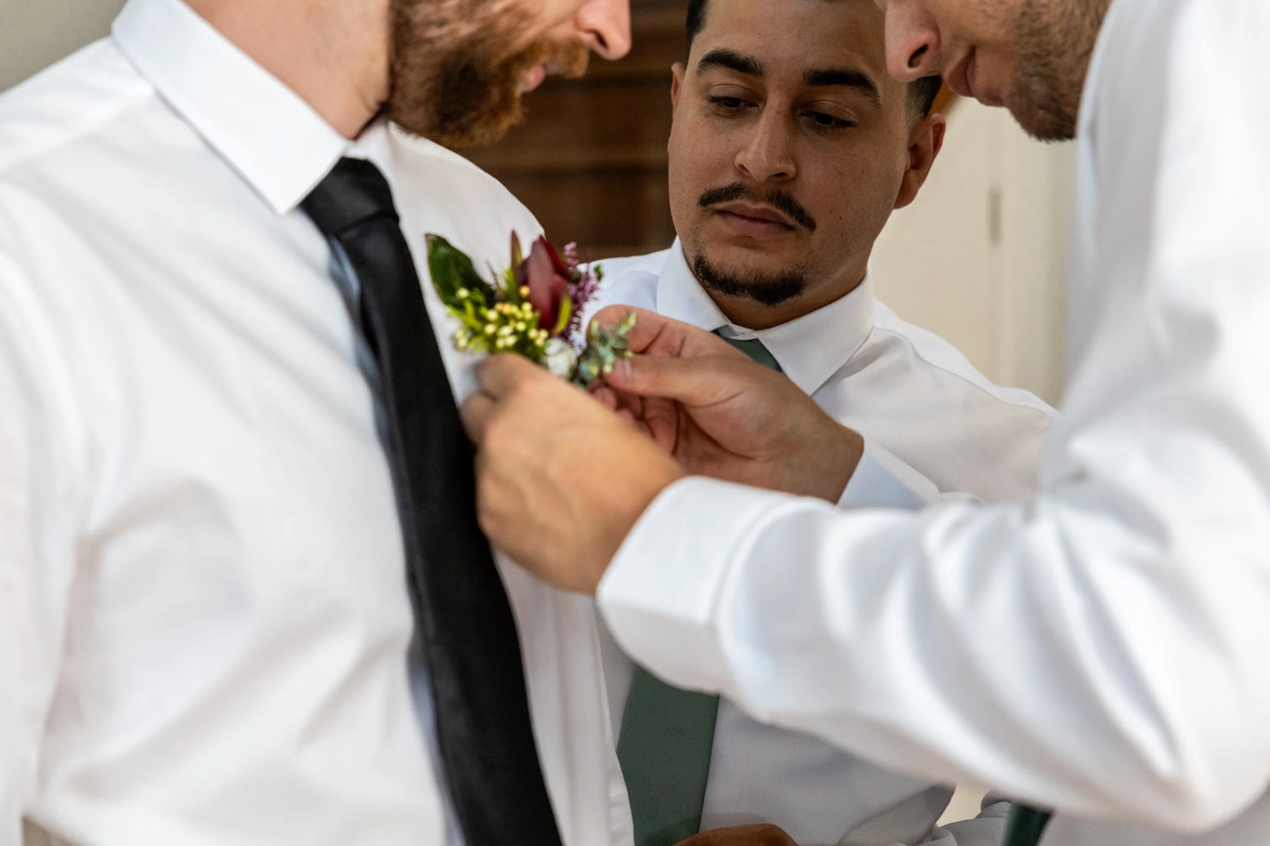 Three men, dressed in white shirts and ties, are helping a man pin a boutonniere onto another man's shirt for a formal occasion.