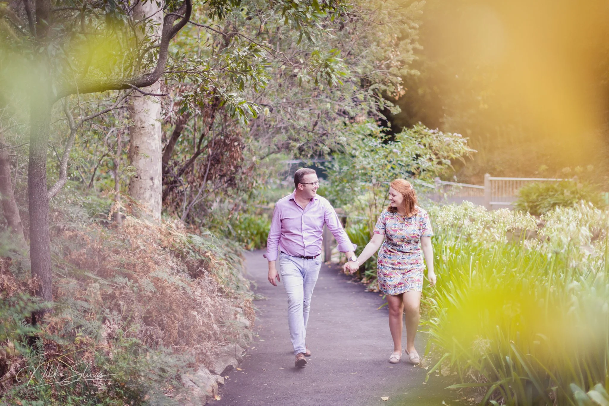 A couple walking hand in hand on a garden pathway, surrounded by trees and flowering plants, with orange and yellow leaves and a fence in the background.