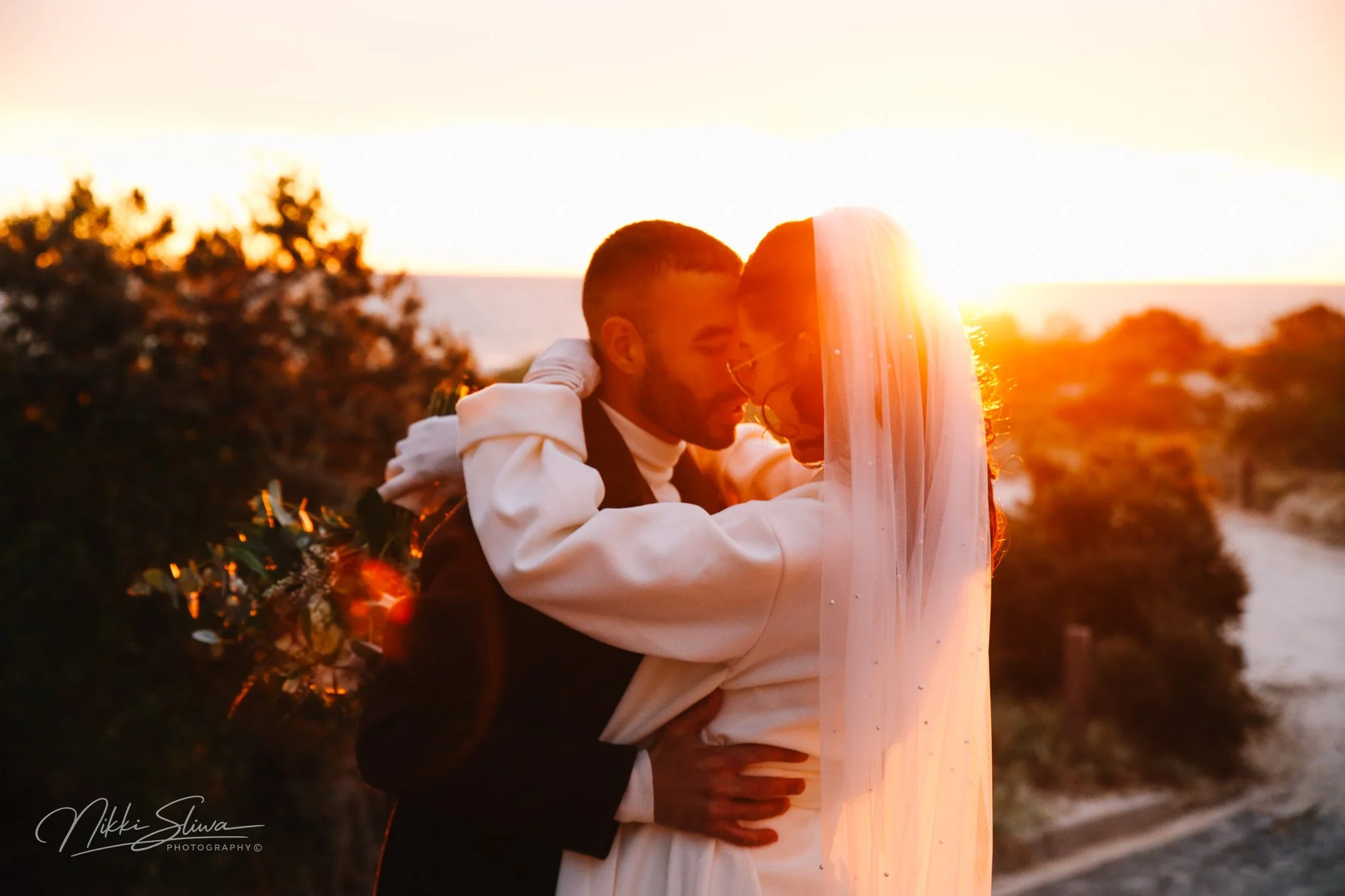 A couple embracing during sunset on a beach, with the sun setting behind them and trees in the background.