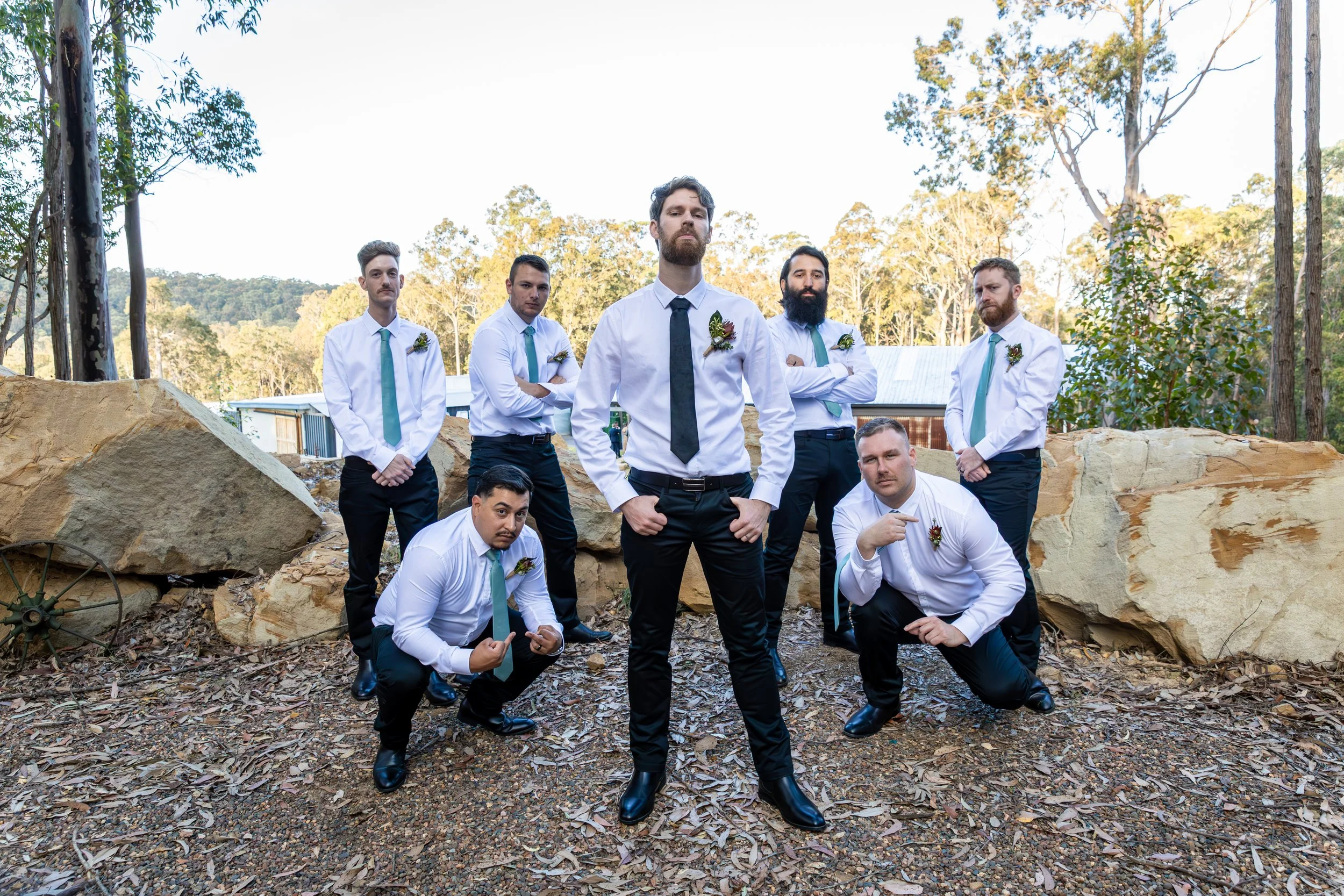 A group of eight men dressed in white shirts, dark pants, and teal ties, standing outdoors with trees and rocks in the background, posing confidently for a photo.