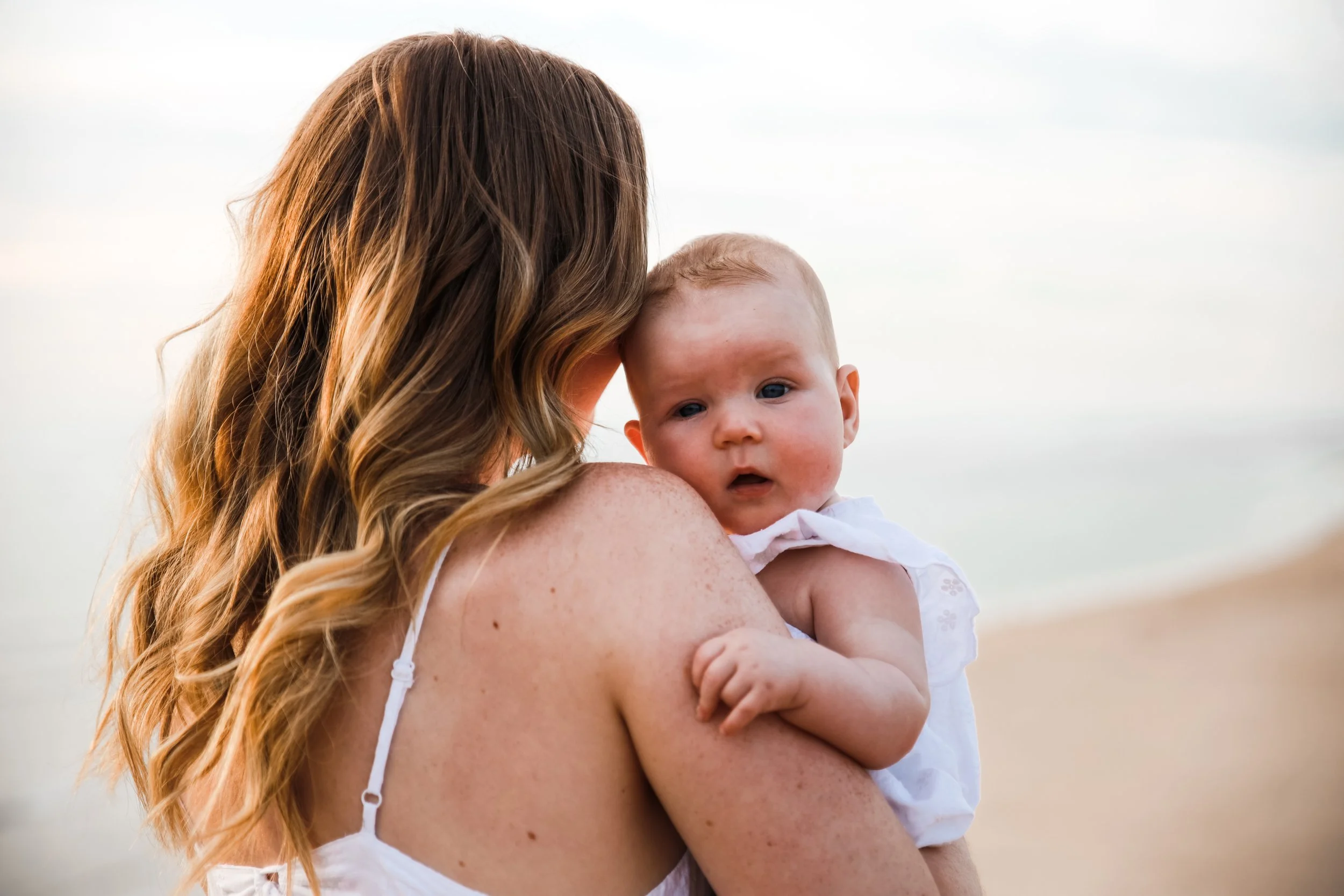 A woman with wavy light brown hair holding a baby with light hair and blue eyes at the beach, with sand and ocean in the background.