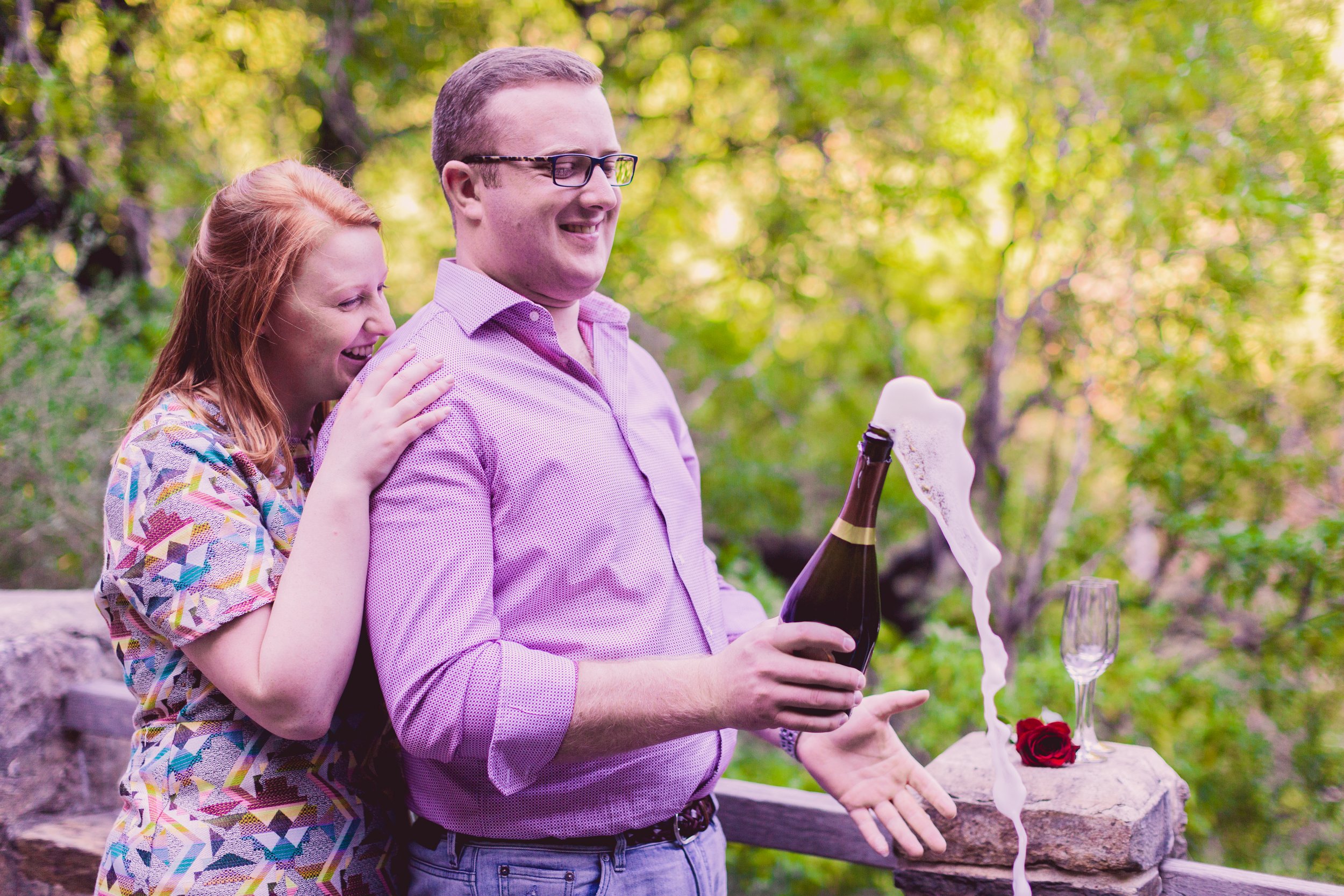 A couple celebrating outdoors with a bottle of champagne, foam spilling out, on a wooden railing, with a glass and a red rose next to it, surrounded by green trees.