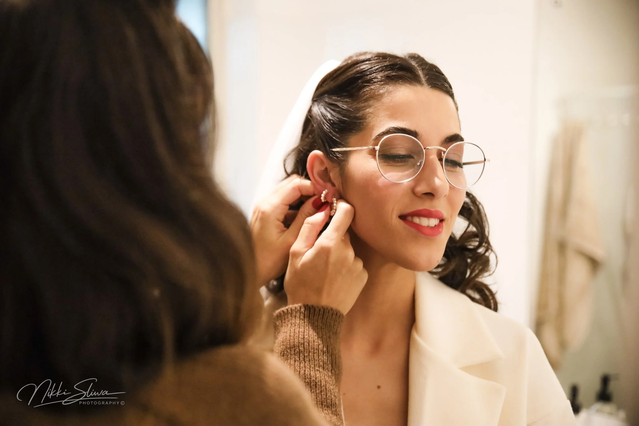 A woman with curly dark hair and glasses is smiling with eyes closed while someone helps her put on earrings, sitting in a white outfit.