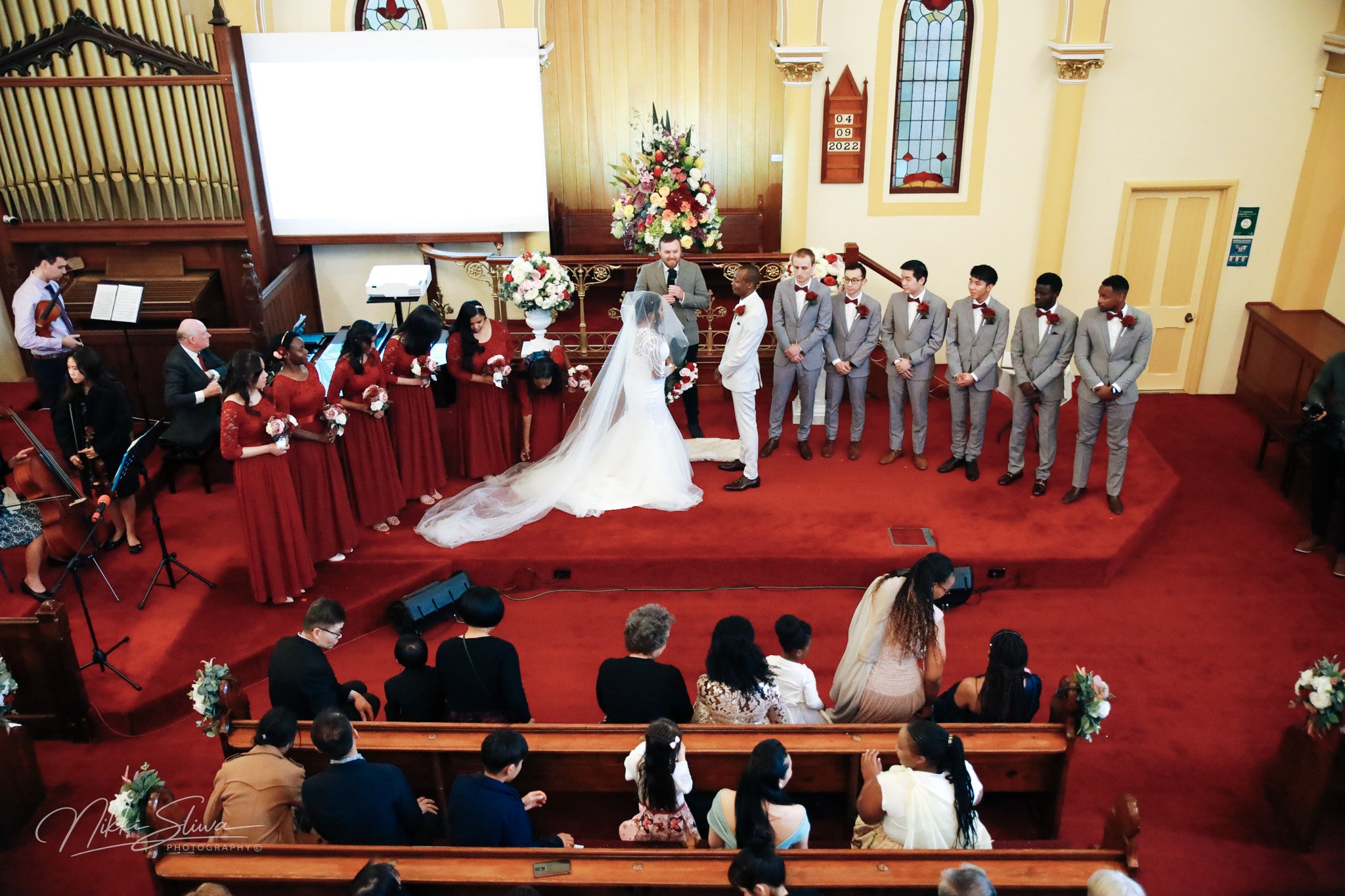 A wedding ceremony taking place in a church with the bride and groom standing at the altar, surrounded by bridesmaids in red dresses and groomsmen in gray suits. Guests are seated and watching the ceremony, and musicians are playing on the side.