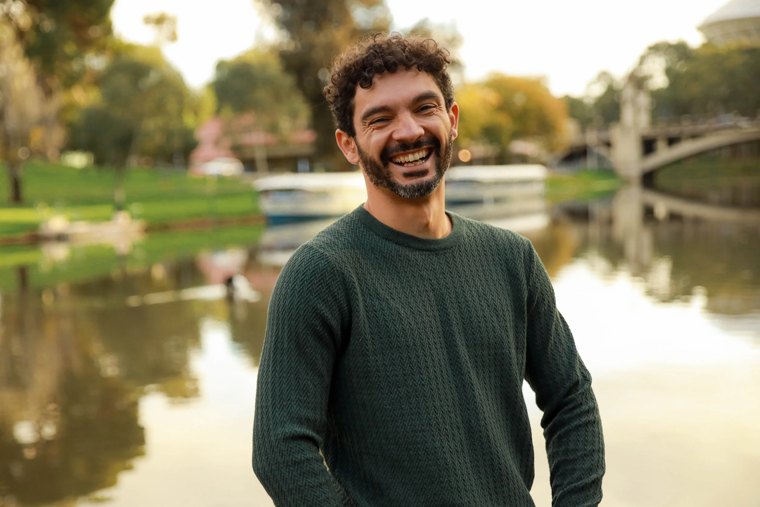 A smiling man with curly hair and a beard standing outdoors near a river. He is wearing a dark green sweater and appears happy. The background shows a park with trees, a bridge, and boats on the water.