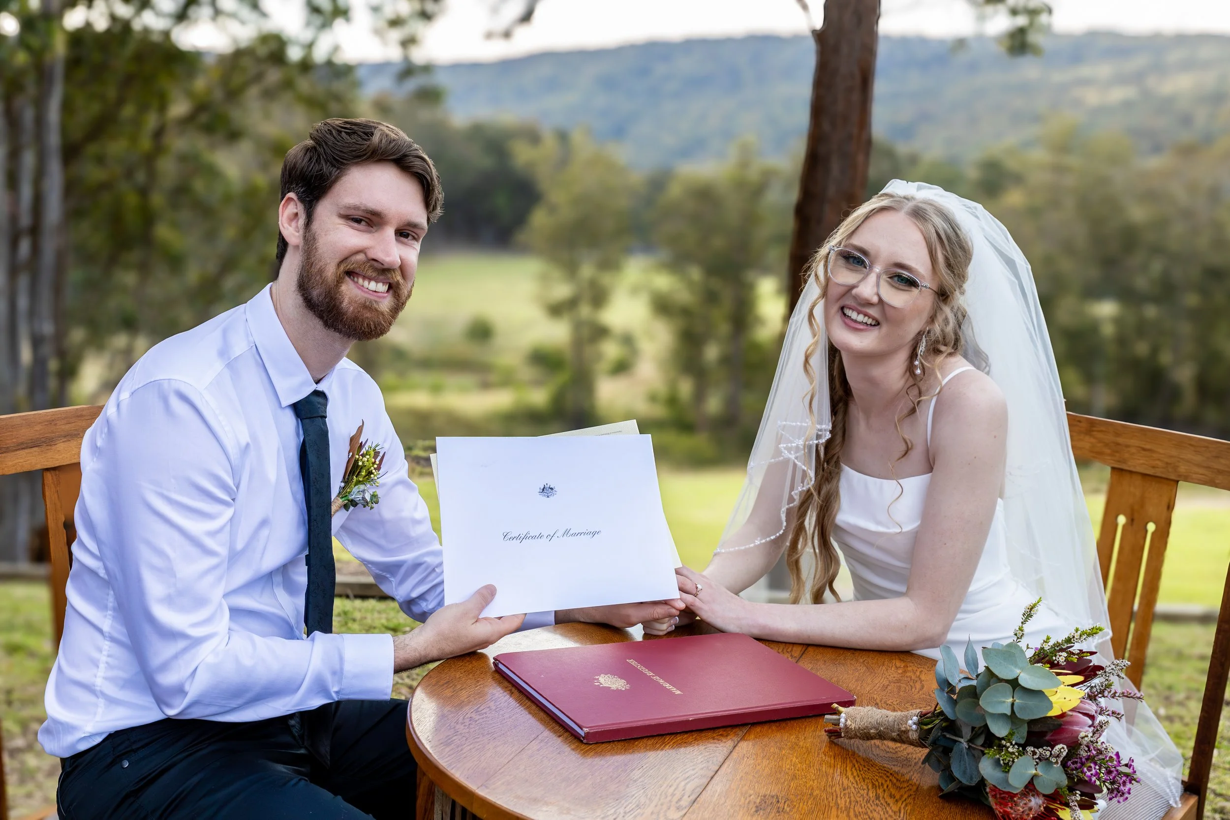 A wedding couple sits outdoors at a wooden table, holding a marriage certificate. The groom has a beard, is dressed in a white shirt with a dark tie, and has a boutonniere. The bride wears glasses, a white wedding dress, and a veil, with a bouquet of