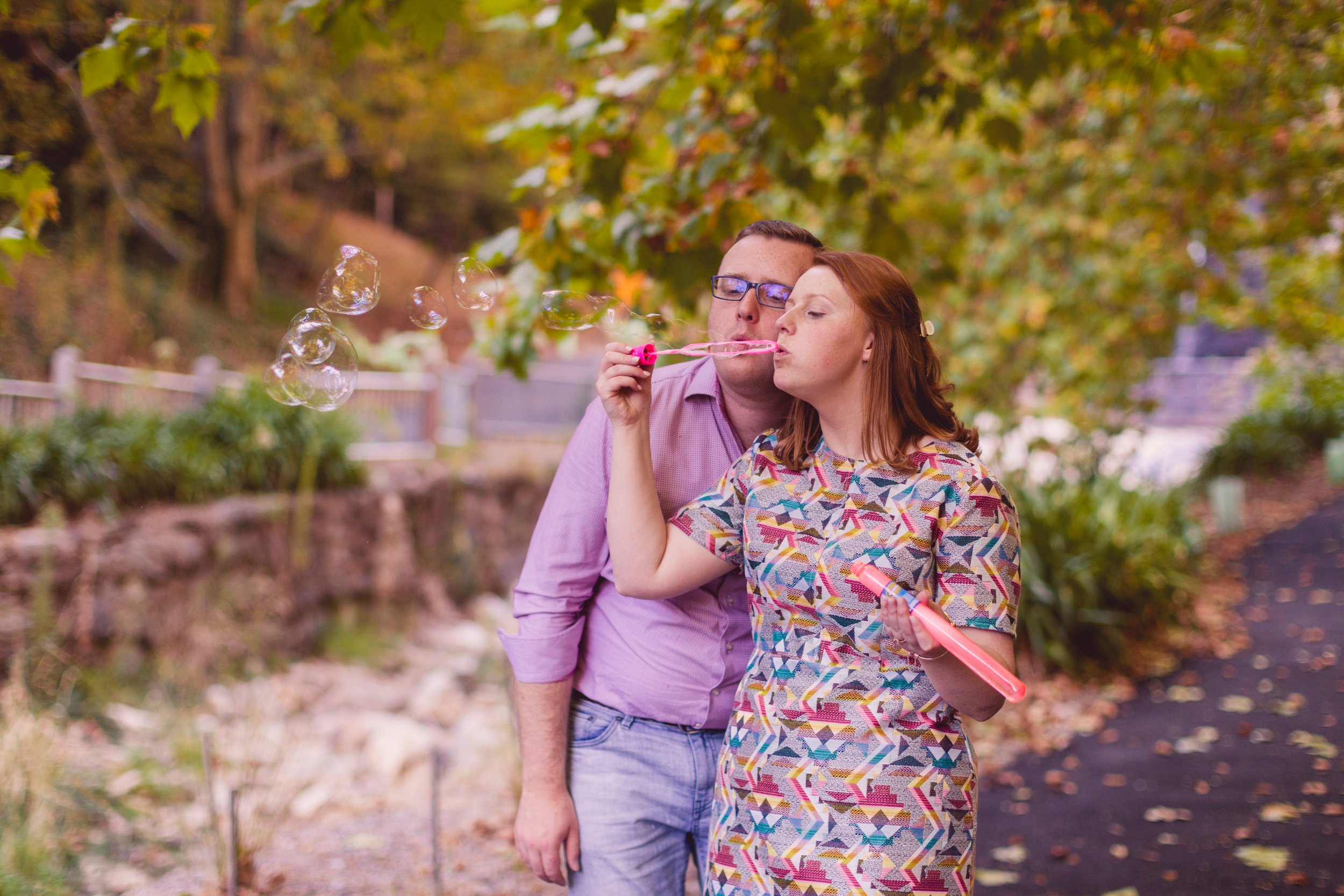 A couple blowing bubbles outdoors in a park with autumn foliage.