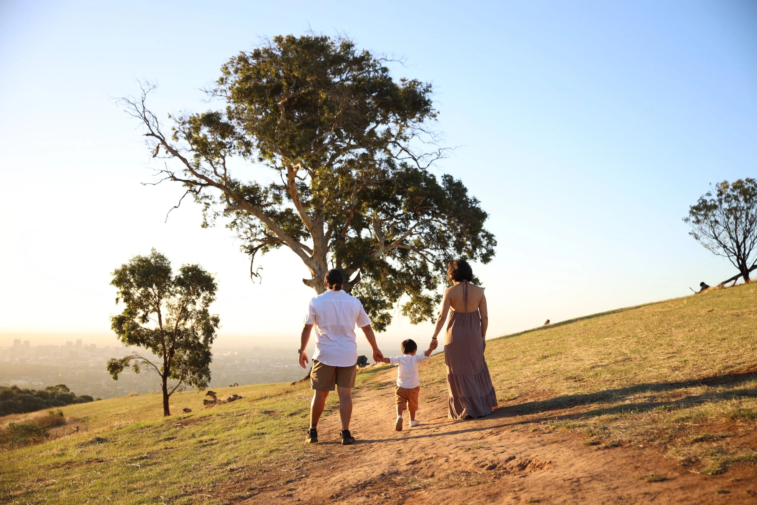 A family of three walking hand in hand on a dirt path in a grassy park during sunset, with trees and a city skyline in the distance.
