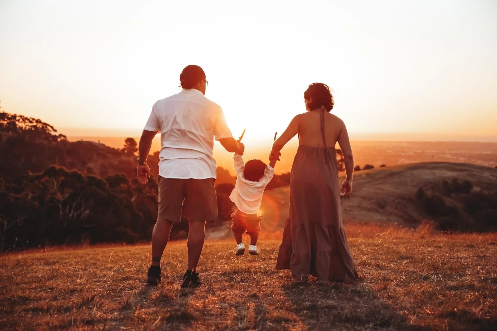 A family of three, a man, woman, and child, holding hands and swinging while walking on a grassy hill at sunset.
