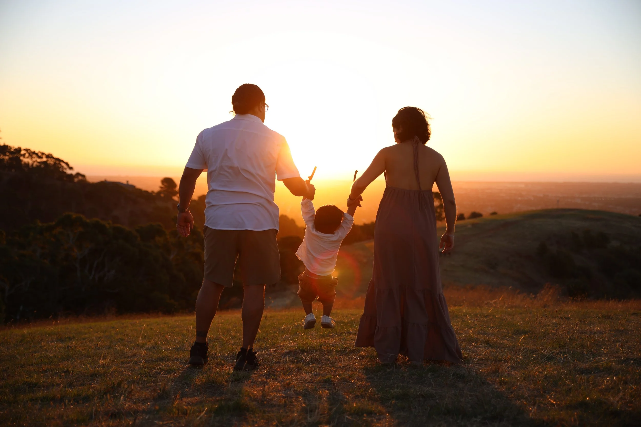 A family of three holding hands and swinging a child between them during sunset on a grassy hill.