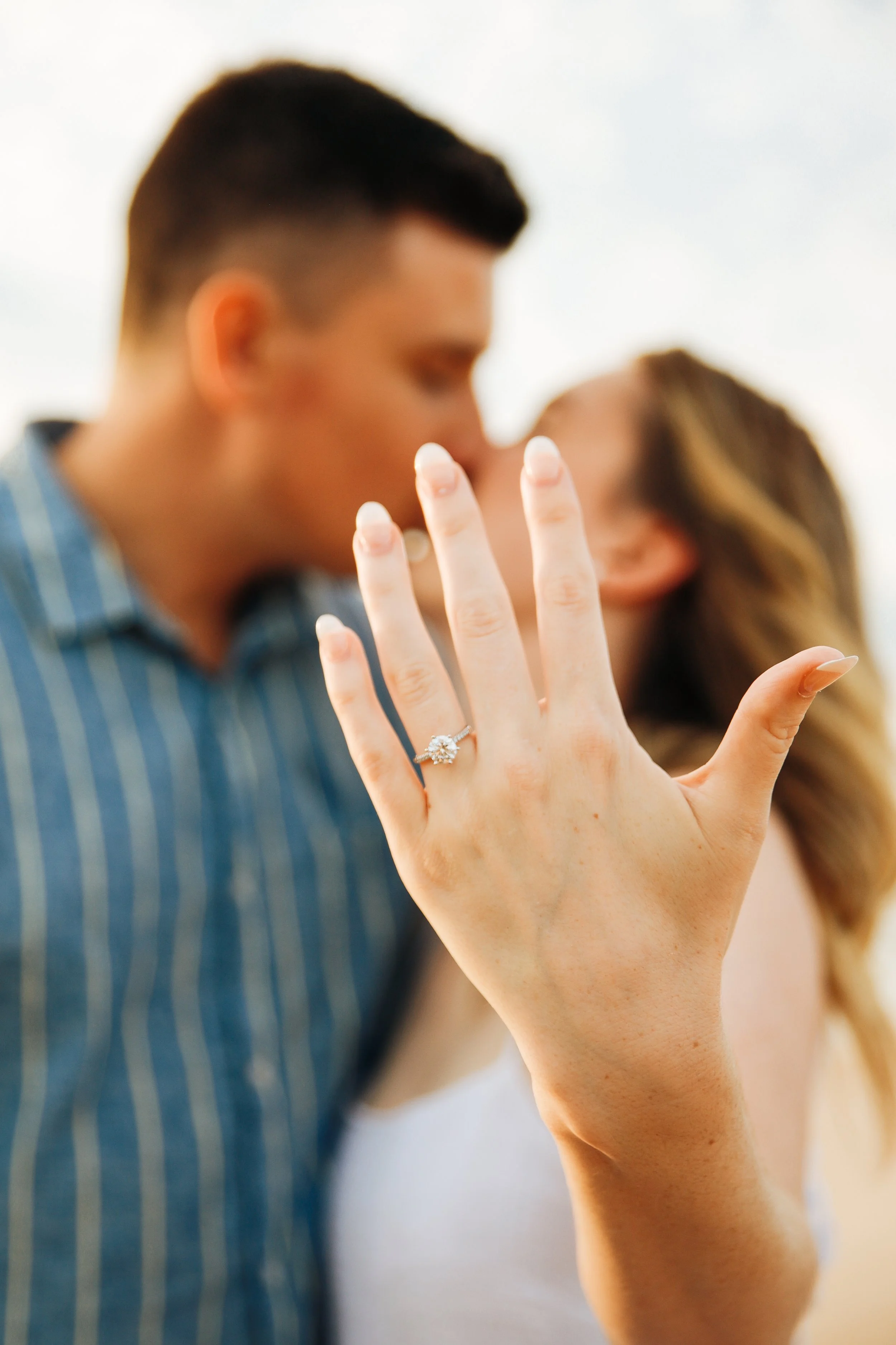 Close-up of a woman showing her left hand with an engagement ring, with a blurred couple kissing in the background.