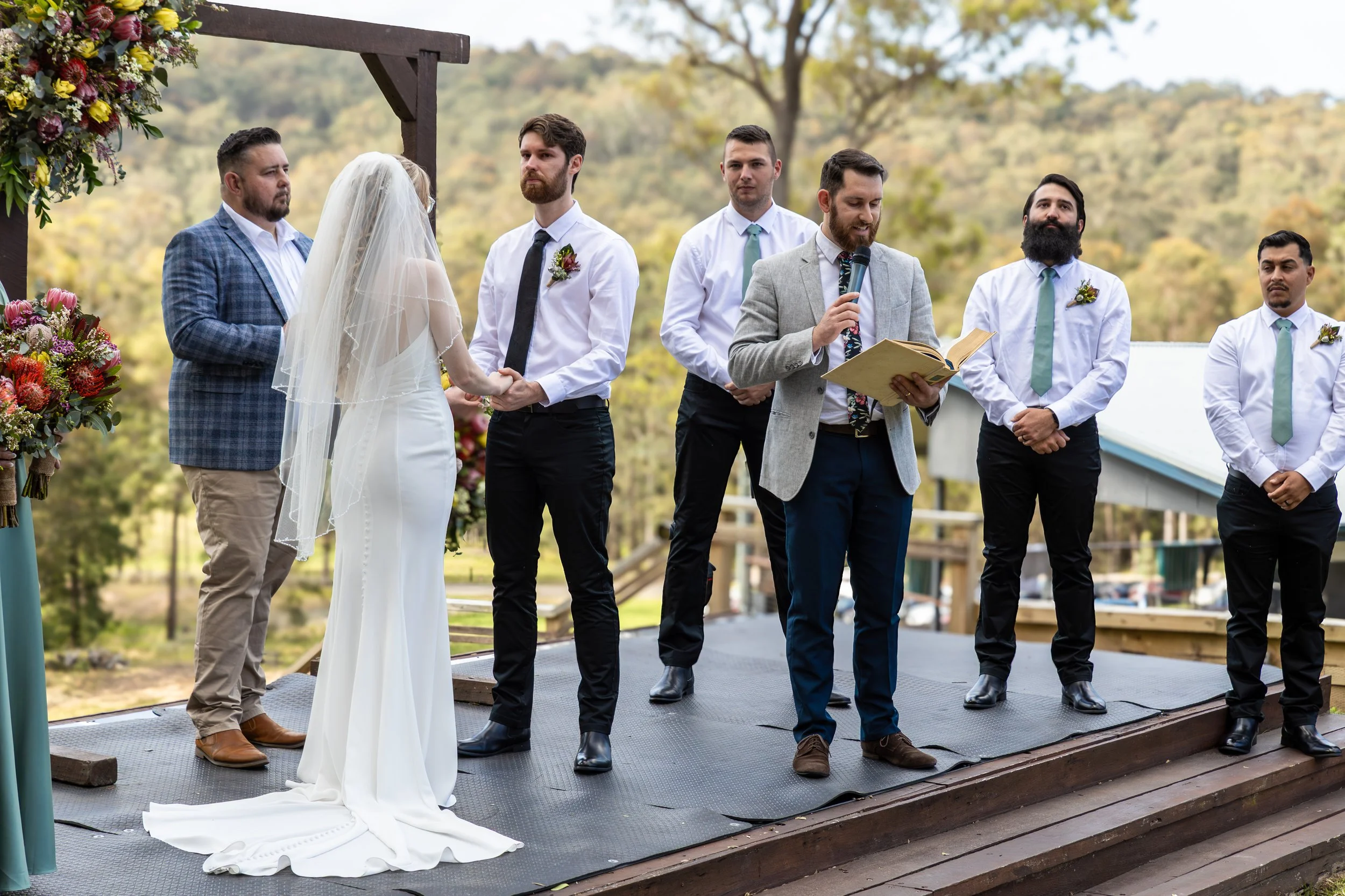 Bride and groom holding hands during an outdoor wedding ceremony with officiant reading from a book, surrounded by groomsmen standing on a wooden platform with trees and hills in the background.