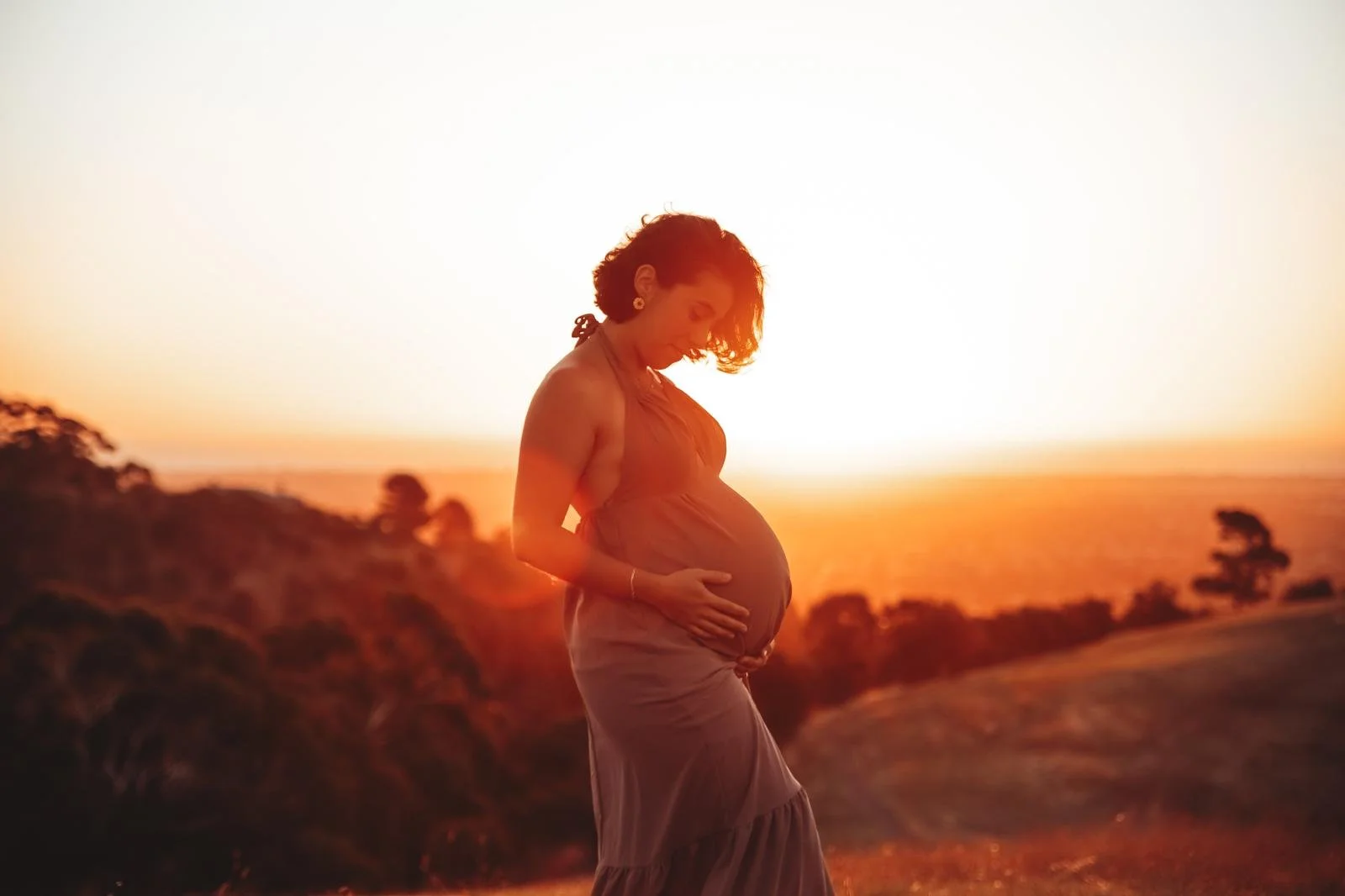 Pregnant woman in a dress standing outdoors during sunset, gently holding her belly.