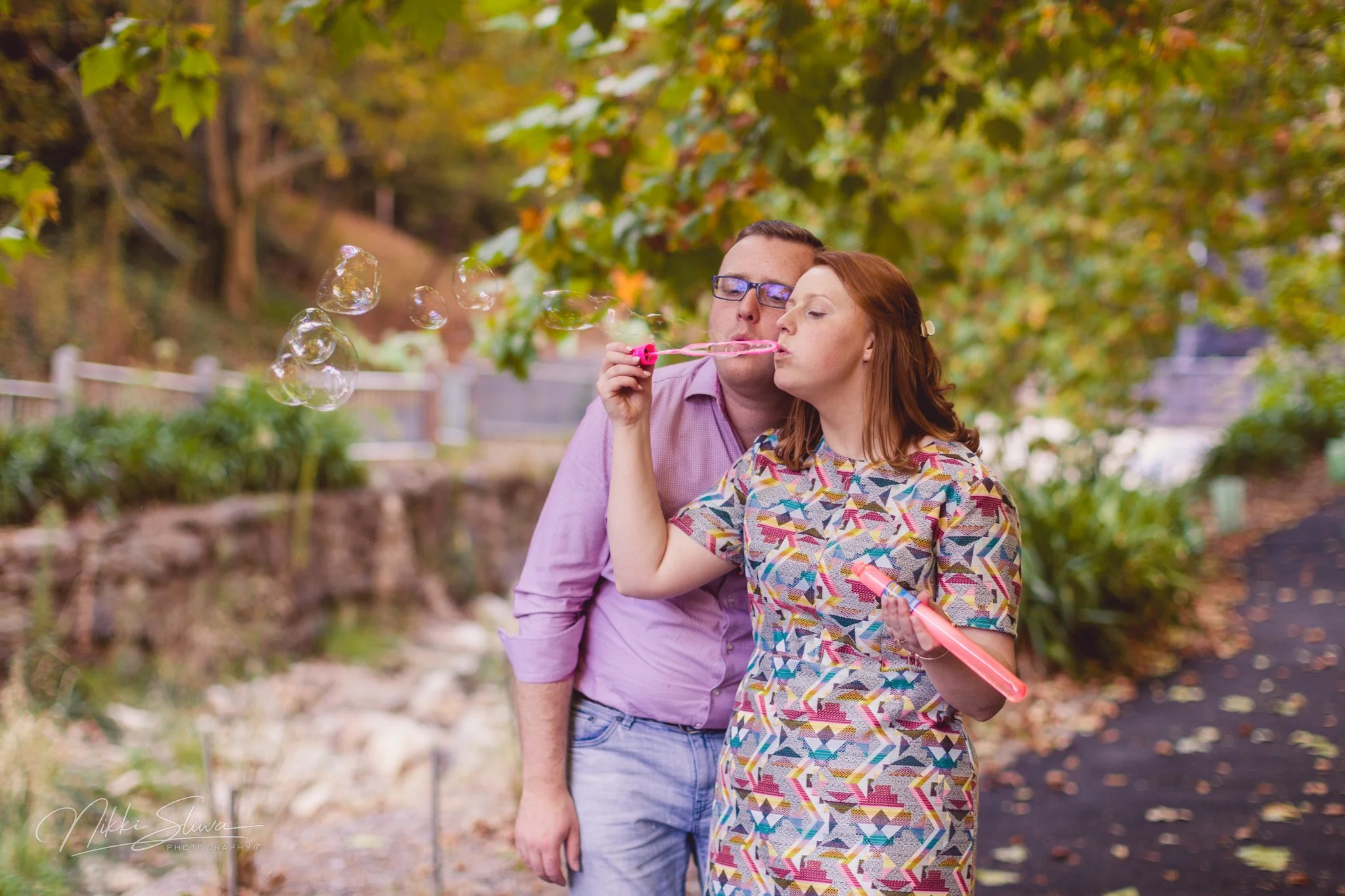A man and woman blowing bubbles outdoors on a fall day with trees and a pathway in the background.
