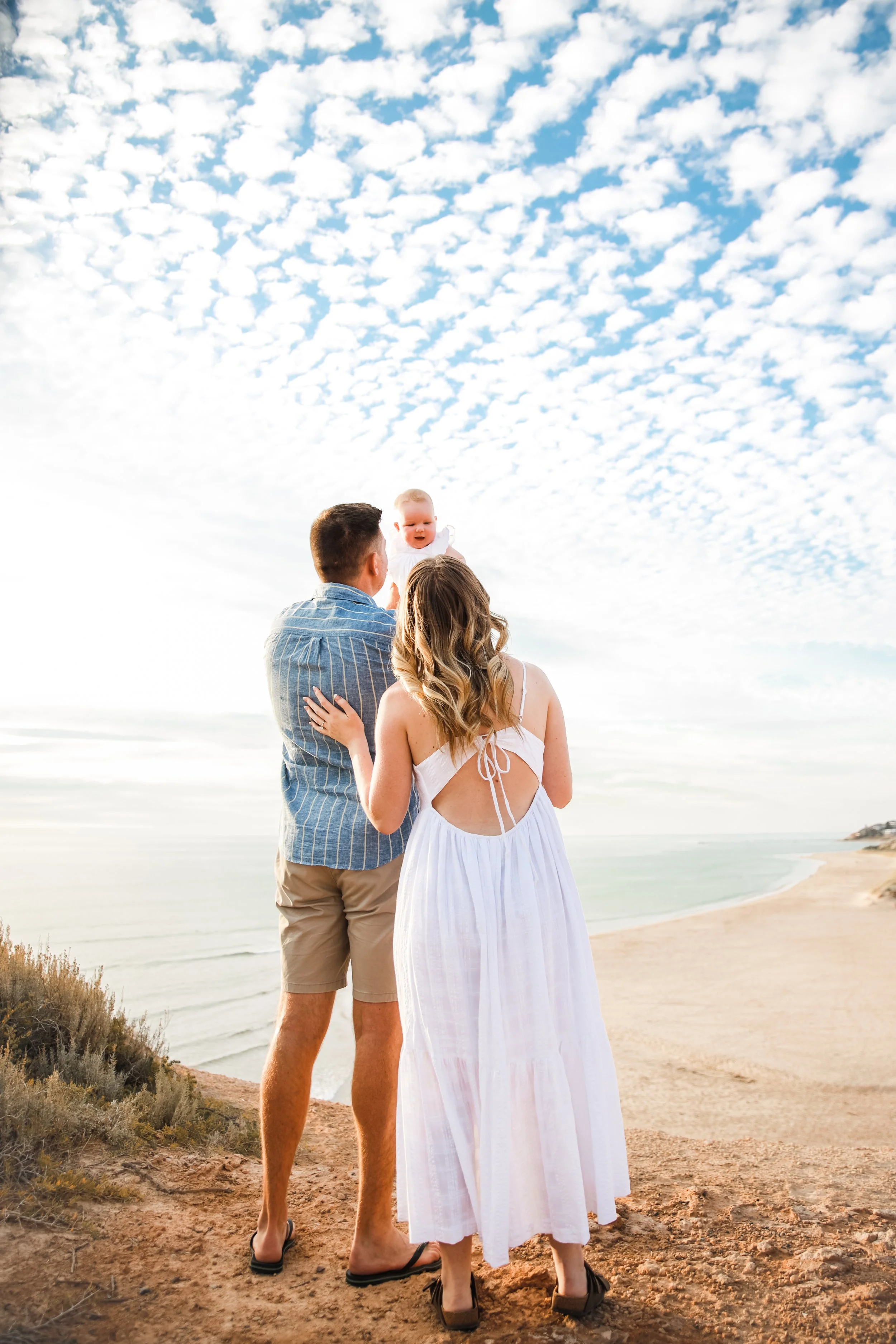 Family of three on a cliff overlooking the ocean, with a cloudy sky above, family standing close together with the father and mother holding their baby.