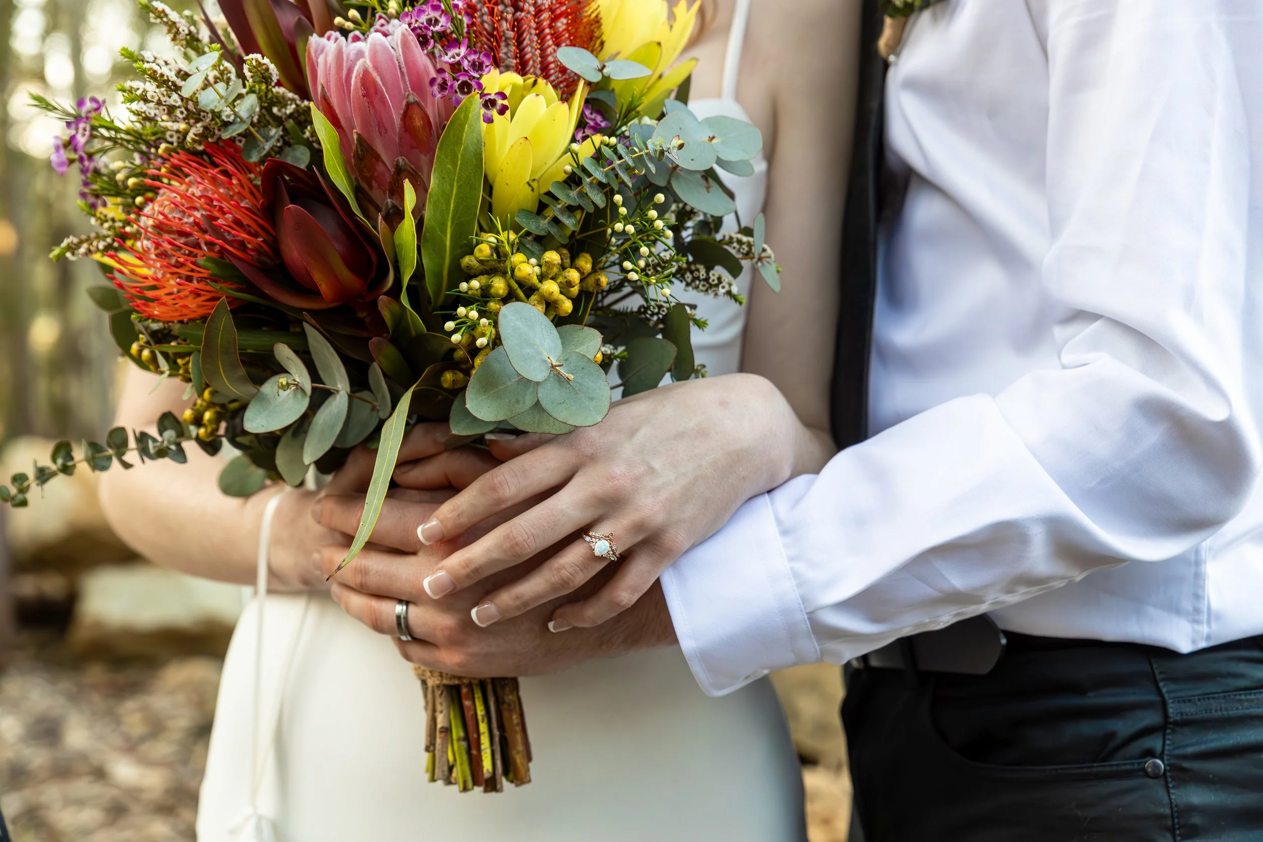 A couple holding a bouquet of colorful flowers, with the woman wearing a wedding ring and a white dress, and the man in a white shirt and black tie, outdoors.