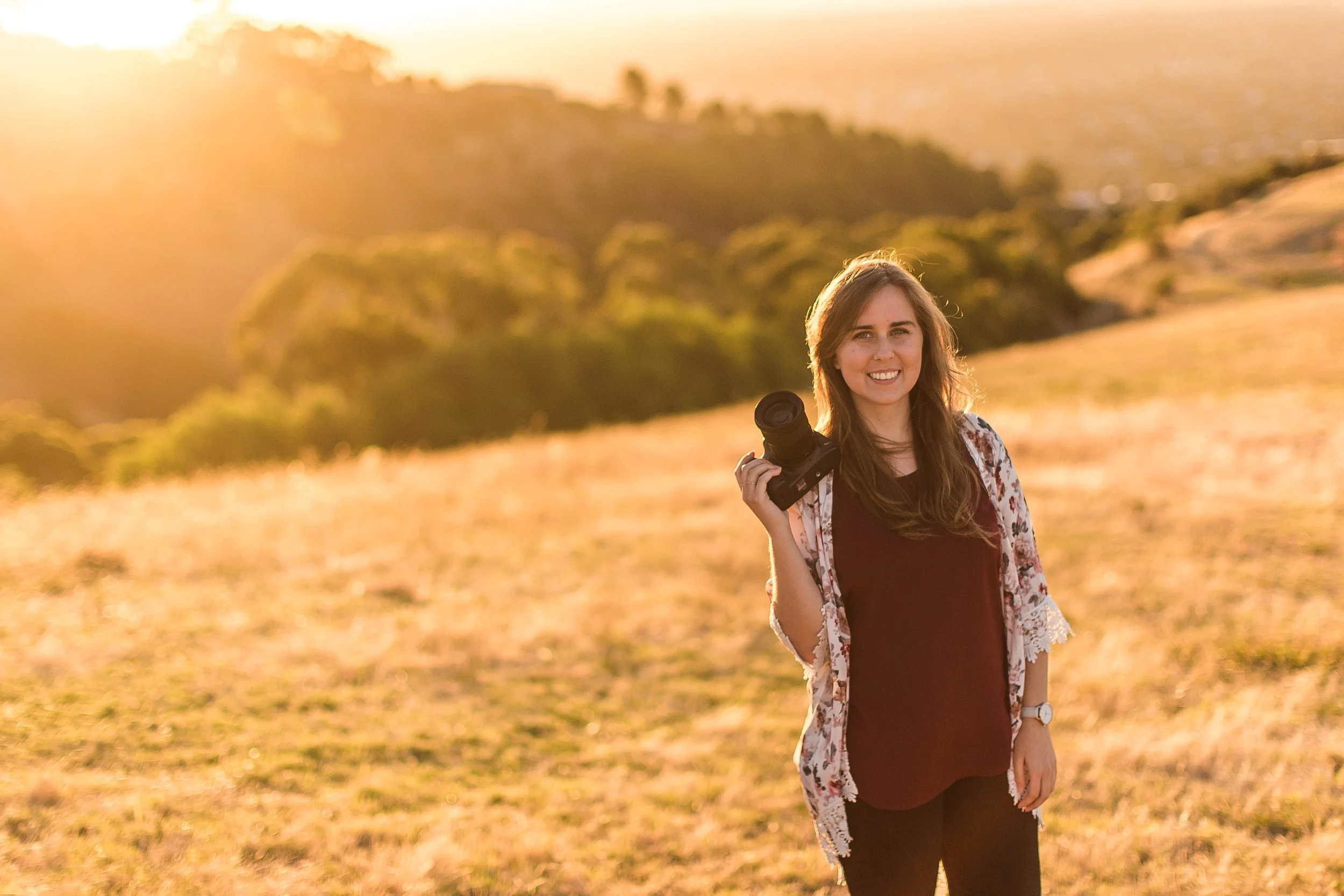 A woman standing in a grassy field at sunset, holding a camera, smiling at the camera.