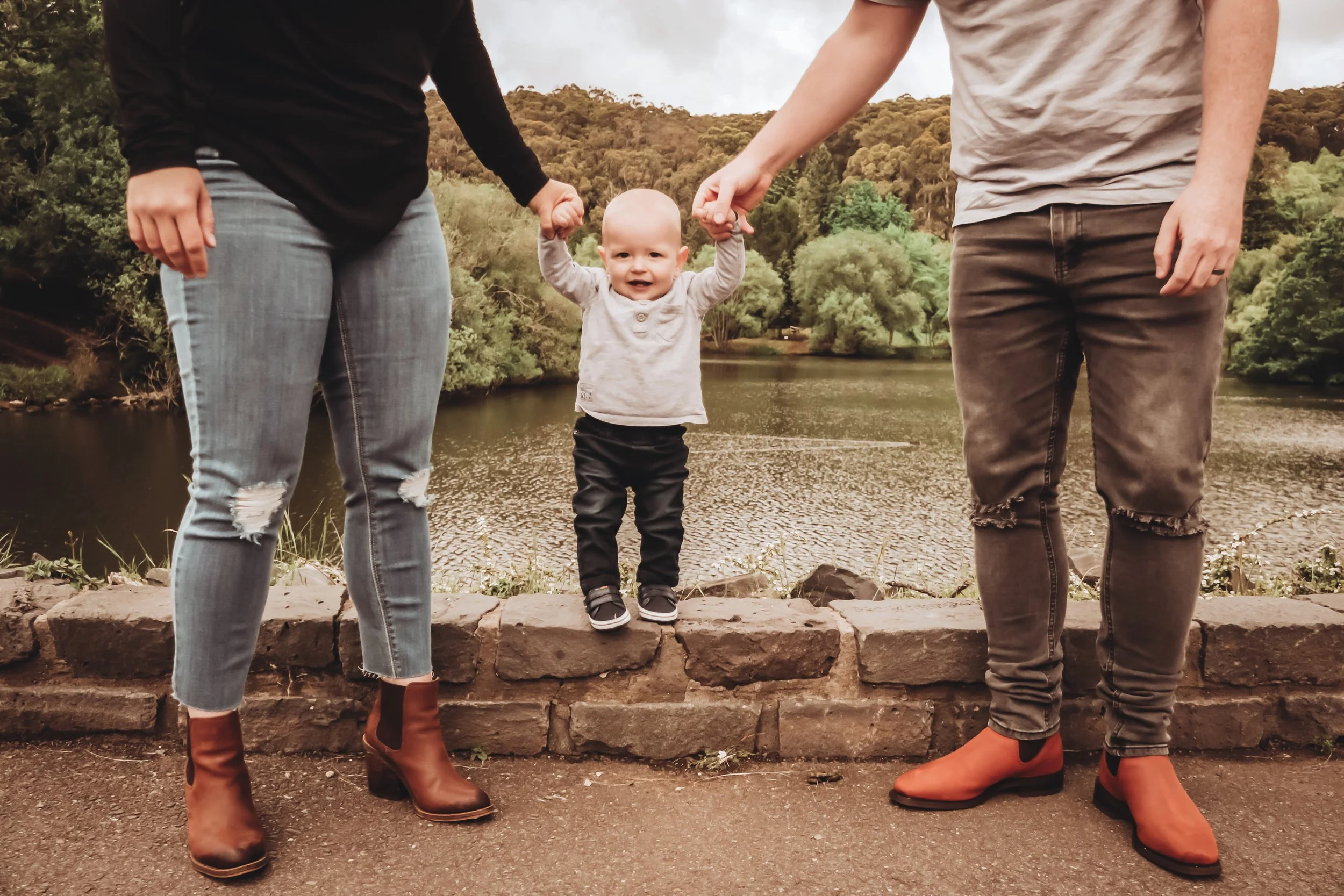 A smiling toddler holding hands with two adults, standing on a stone ledge with a lake and trees in the background.