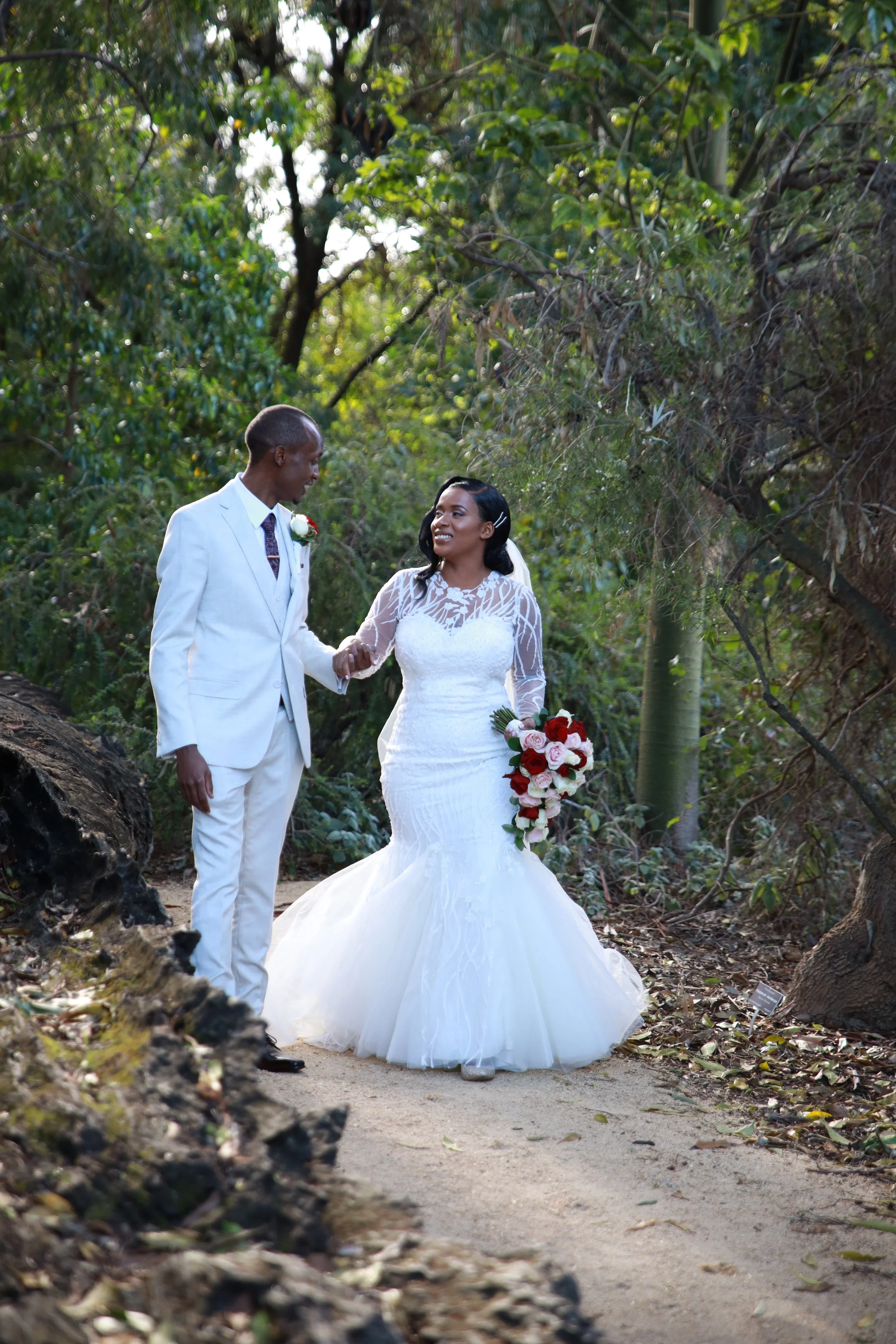 A bride and groom holding hands and smiling in a wooded area during their wedding photo, with the bride holding a bouquet of red and white flowers.