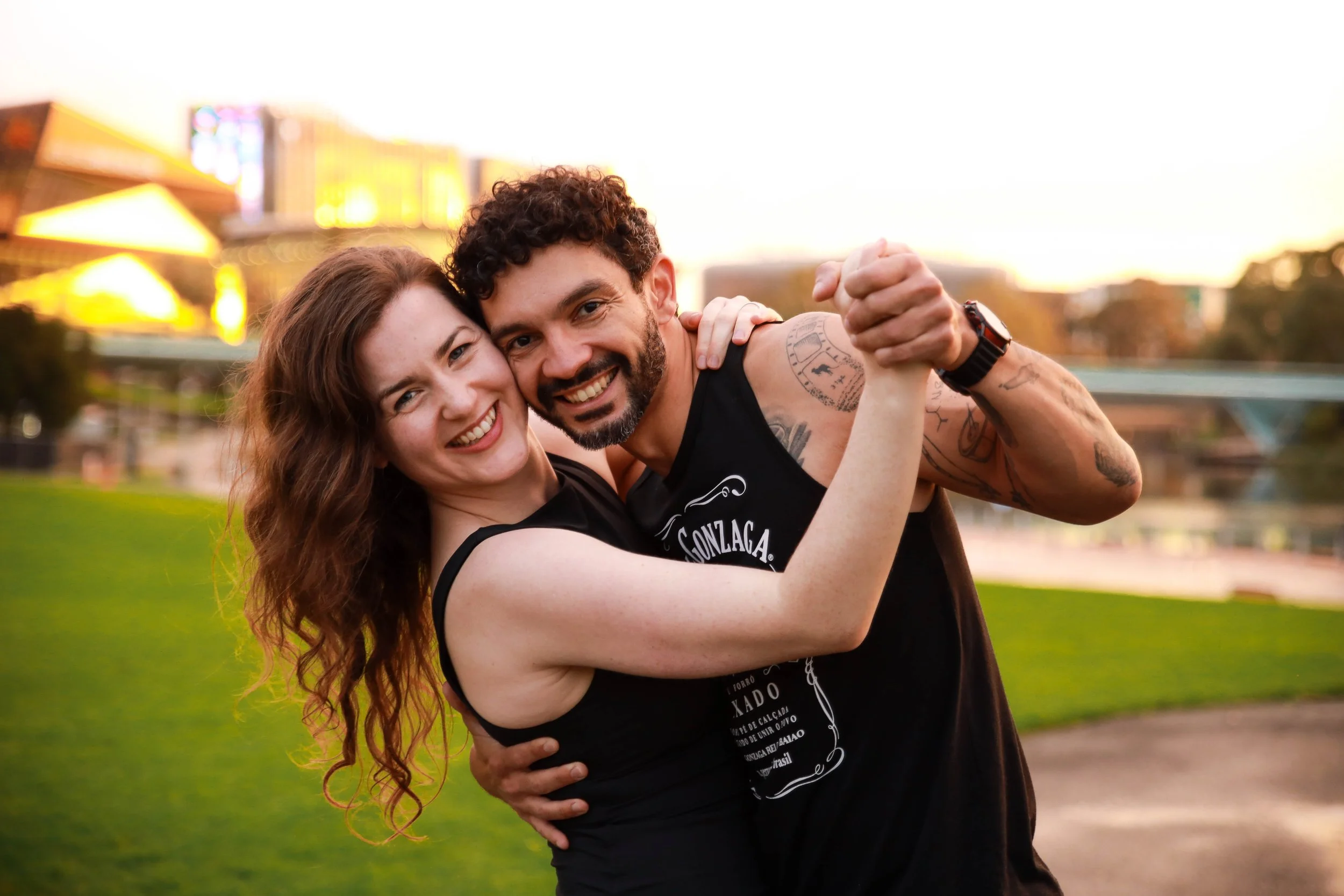 A smiling couple with the woman in a black tank top and the man in a black sleeveless shirt, embracing and holding hands in a dance pose outdoors on a grassy field during sunset.