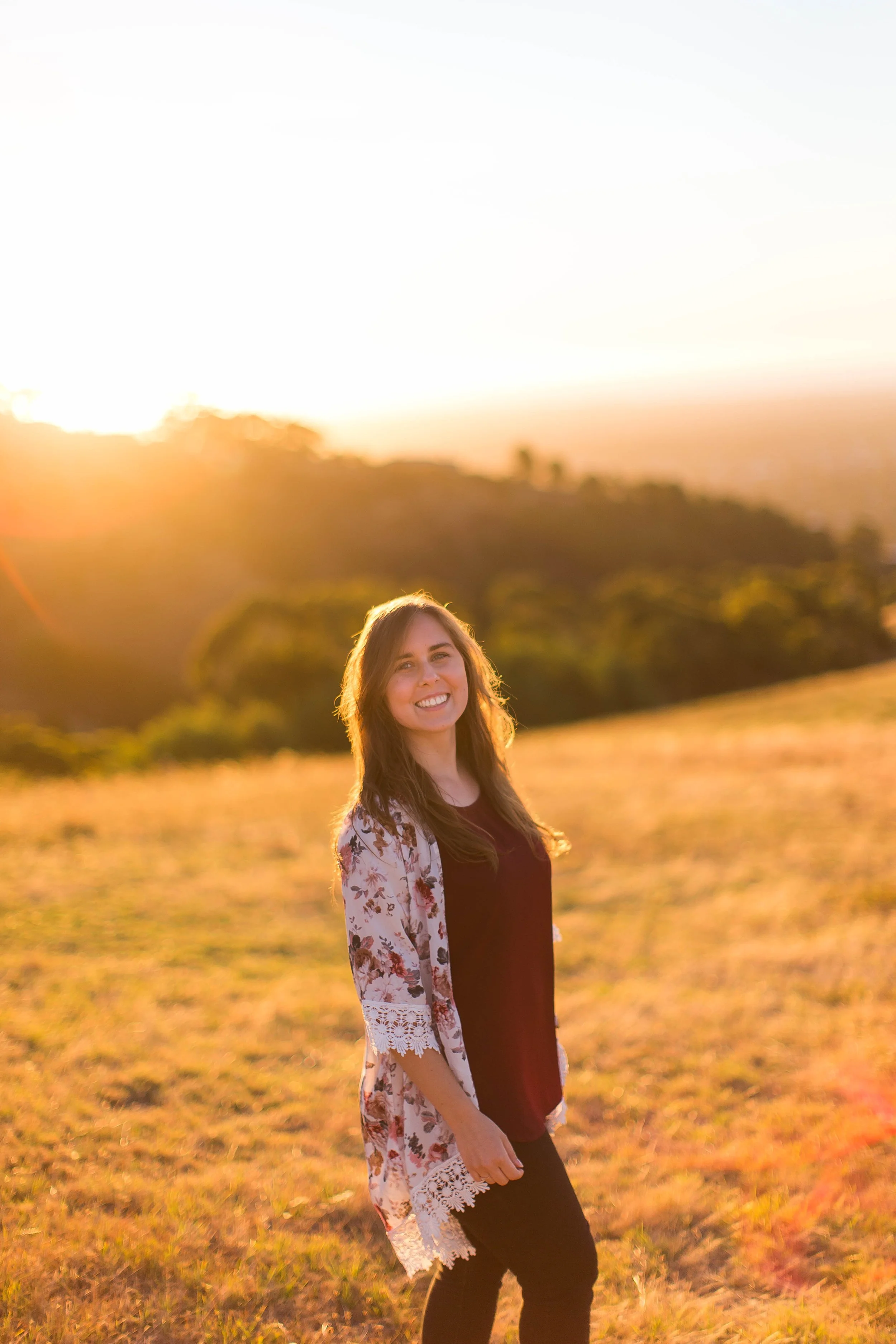 A smiling woman standing outdoors in a grassy field during sunset, with trees and hills in the background.