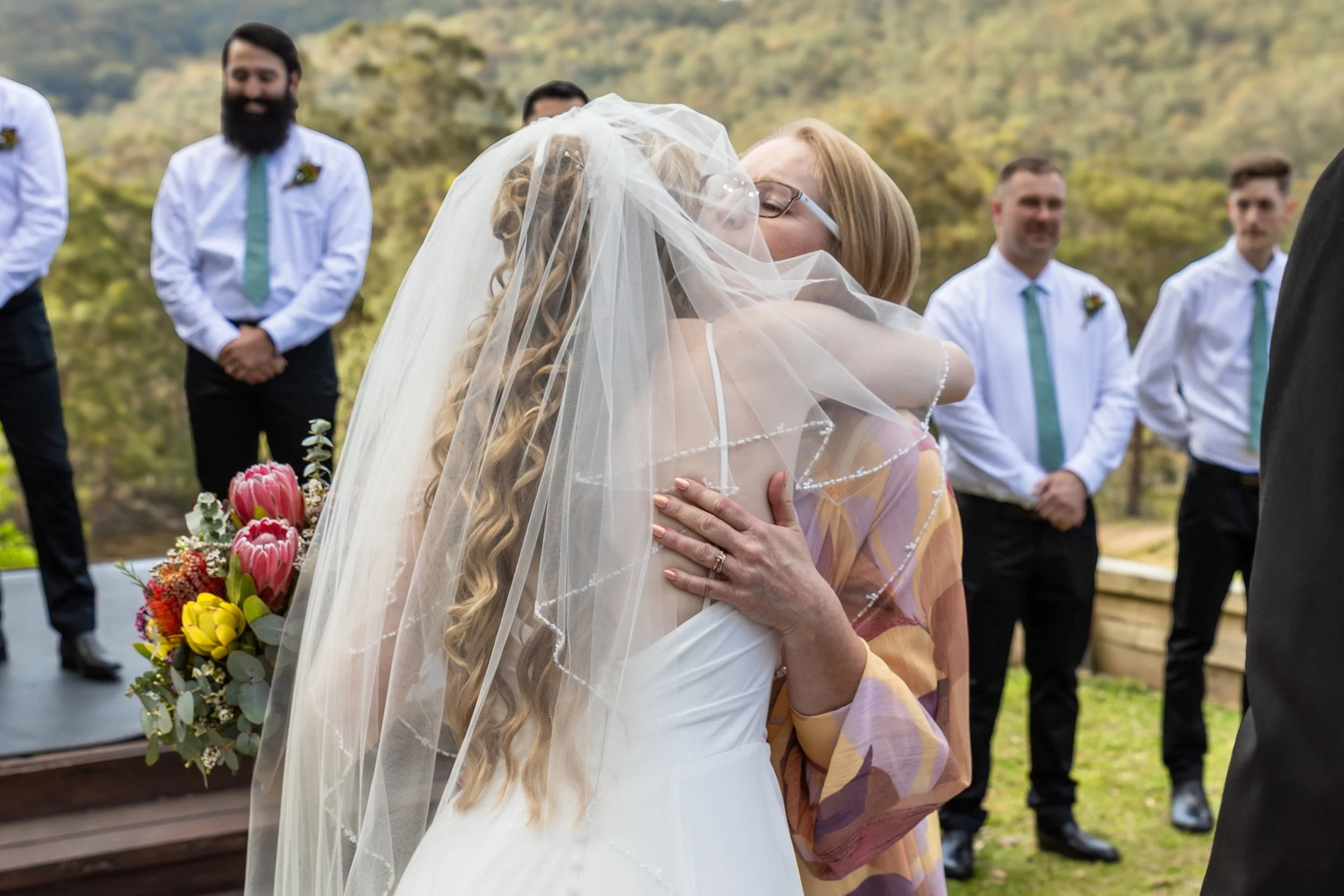 A bride in a white wedding dress and veil hugging a woman outdoors during a wedding ceremony, with men in white shirts and green ties in the background.