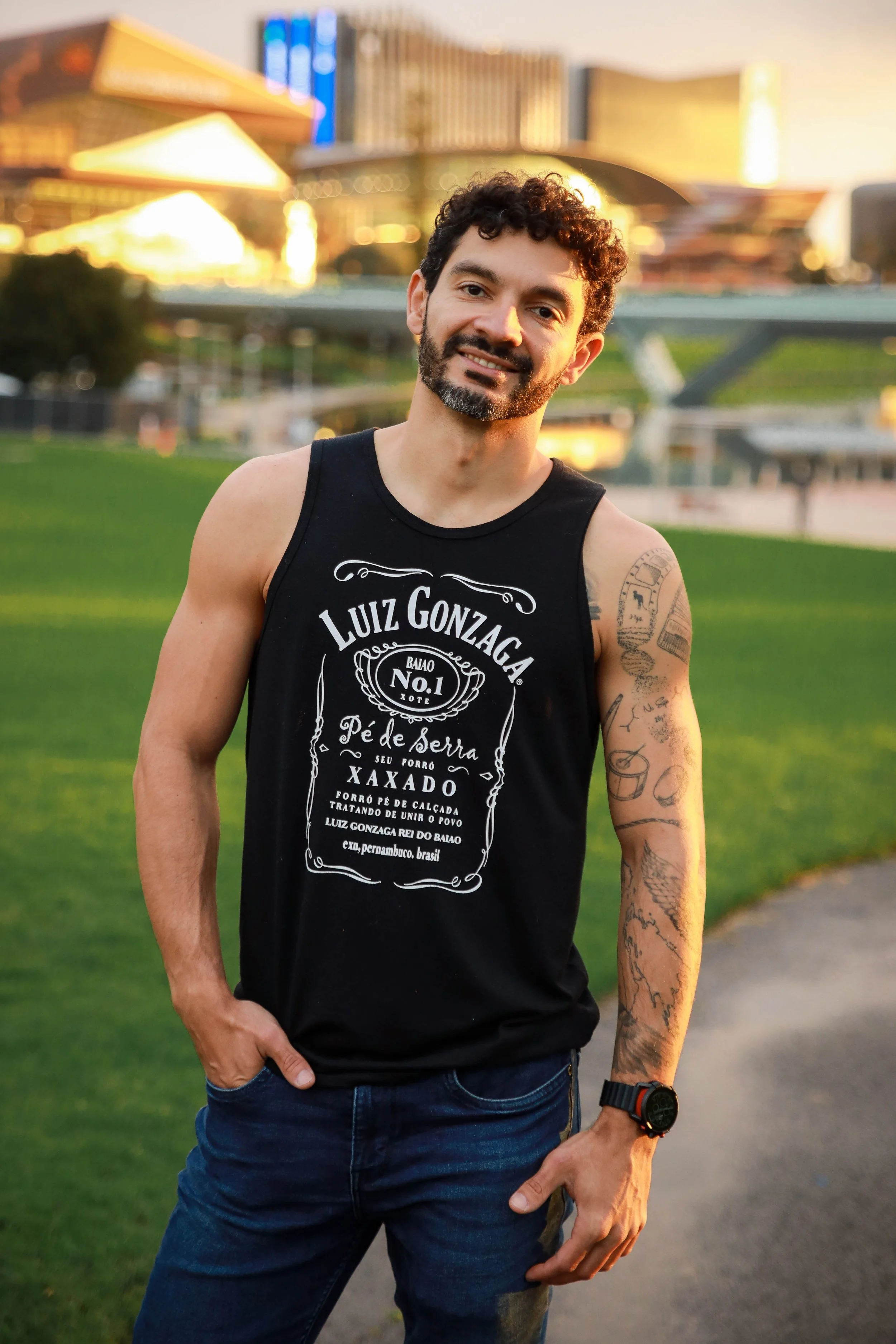 A man with curly hair, a beard, and tattoos on his left arm is smiling and standing outdoors on a grassy area with a modern architectural background during sunset. He is wearing a black sleeveless shirt with white text and dark blue jeans.