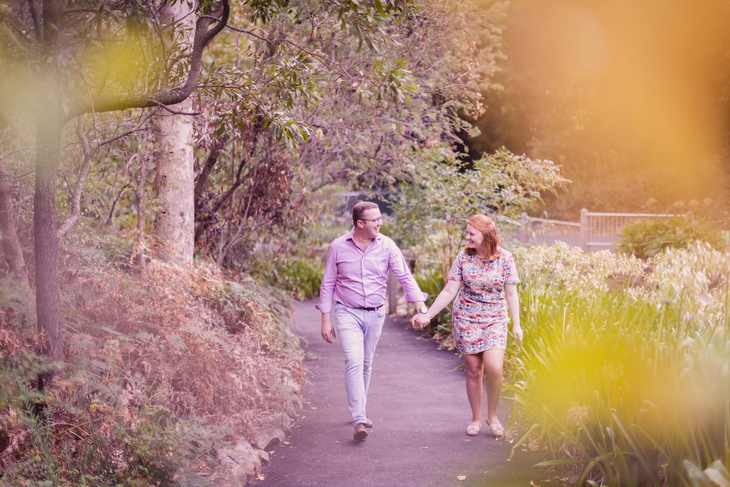 A man and woman walking hand in hand along a garden path, smiling and looking at each other, surrounded by trees and flowering plants.