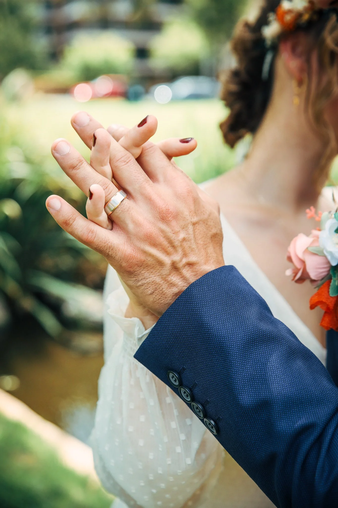Close-up of two hands holding each other during a wedding, with the bride's hand showing a wedding band and dark red nail polish, and the groom's hand in a blue suit sleeve. The bride has a floral headpiece and a white dress, with blurred greenery an