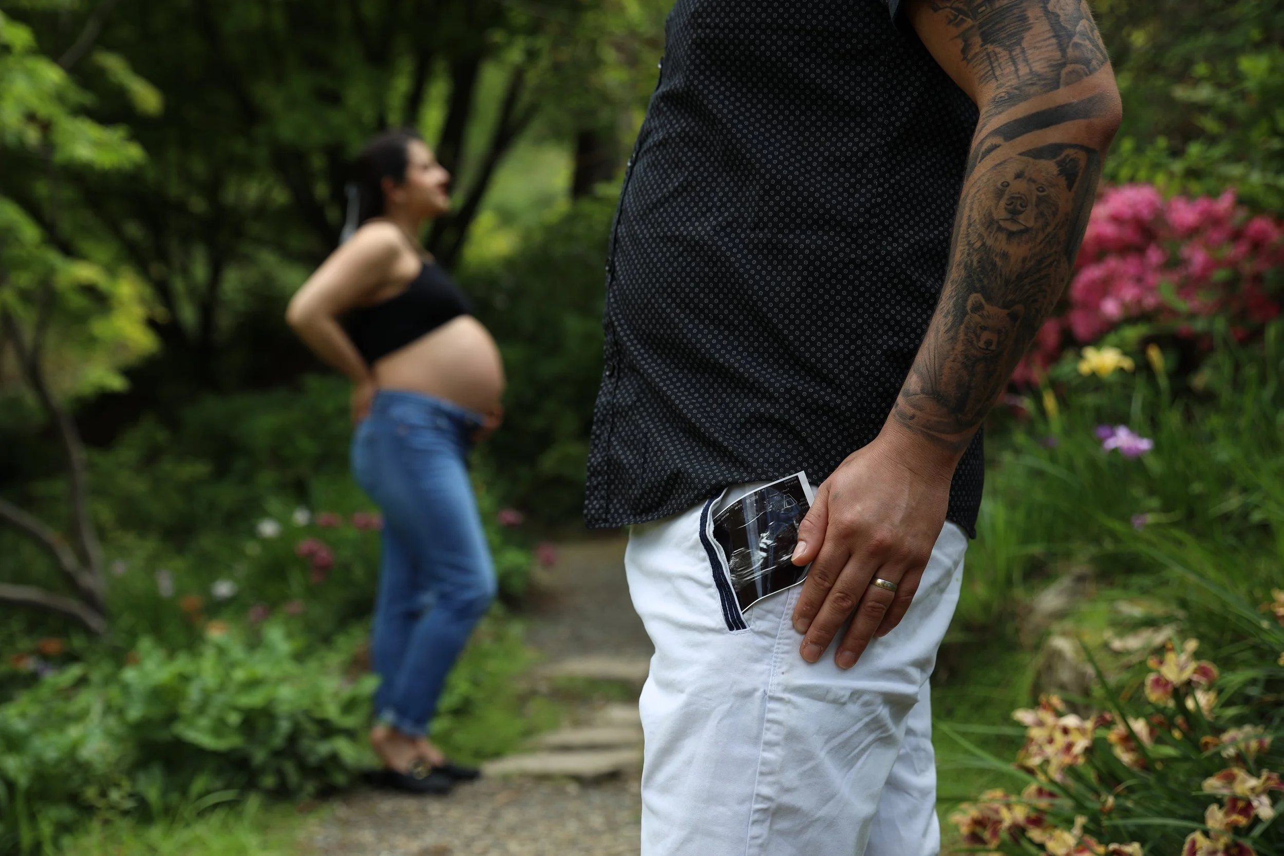 A man with tattoos in white pants is holding a pregnancy test in his pocket while standing in front of a pregnant woman in a garden.